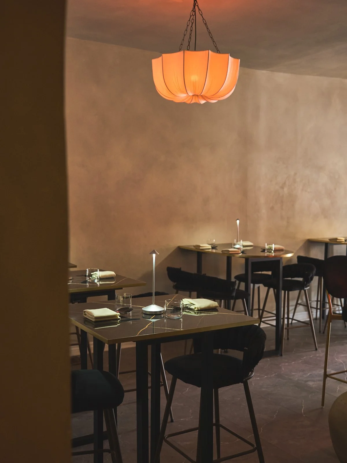 A dimly lit restaurant with a large peach-colored pendant lamp hanging from the ceiling, several tables set with glassware, napkins, and candles, and black chairs. Kozhi Restaurant. Brand Identity