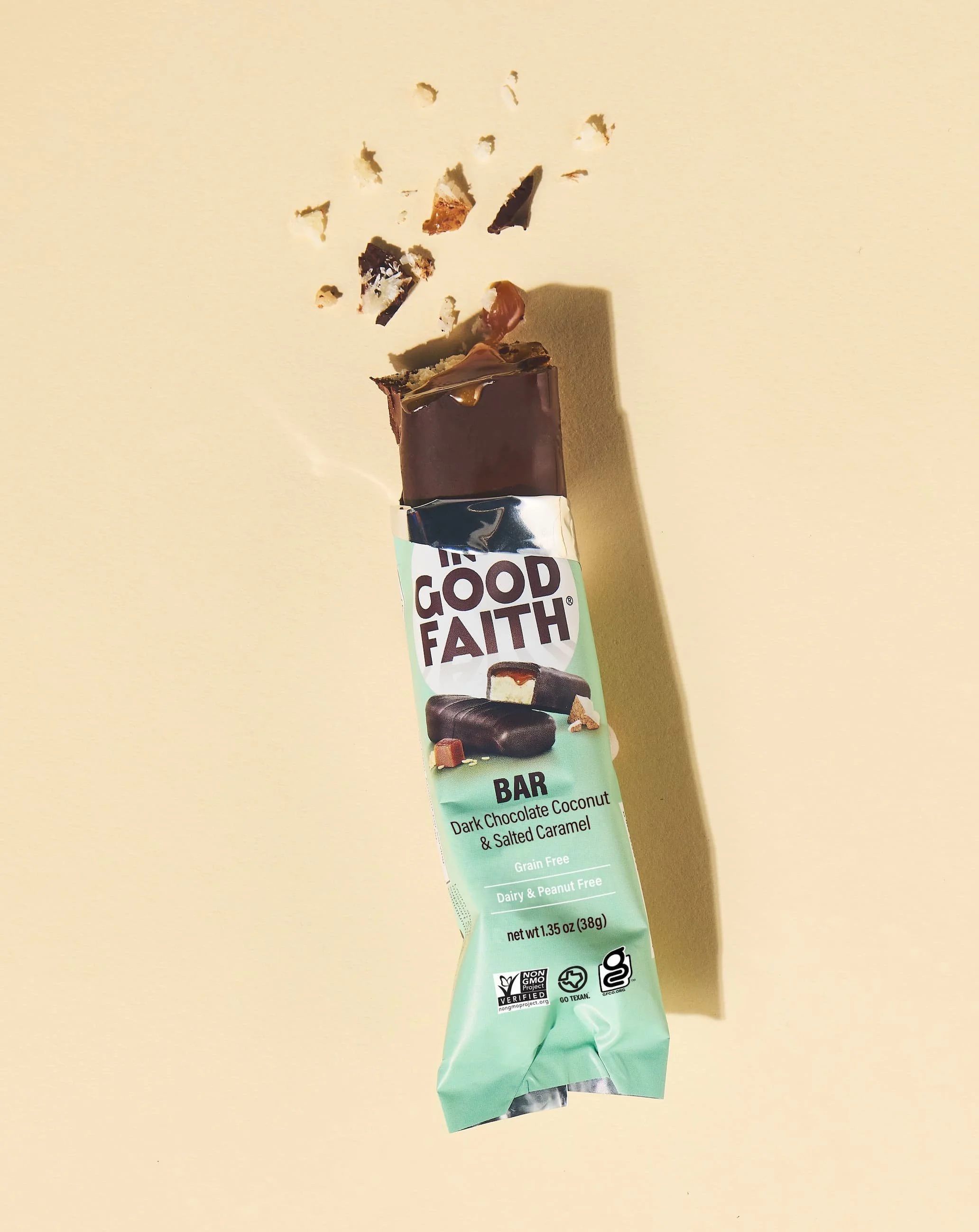 In Good Faith chocolate bar wrapper with broken pieces of dark chocolate, coconut, and salted caramel falling out, on a beige surface. Photographed by Nick Ghattas, food stylist is Gumption Studio.