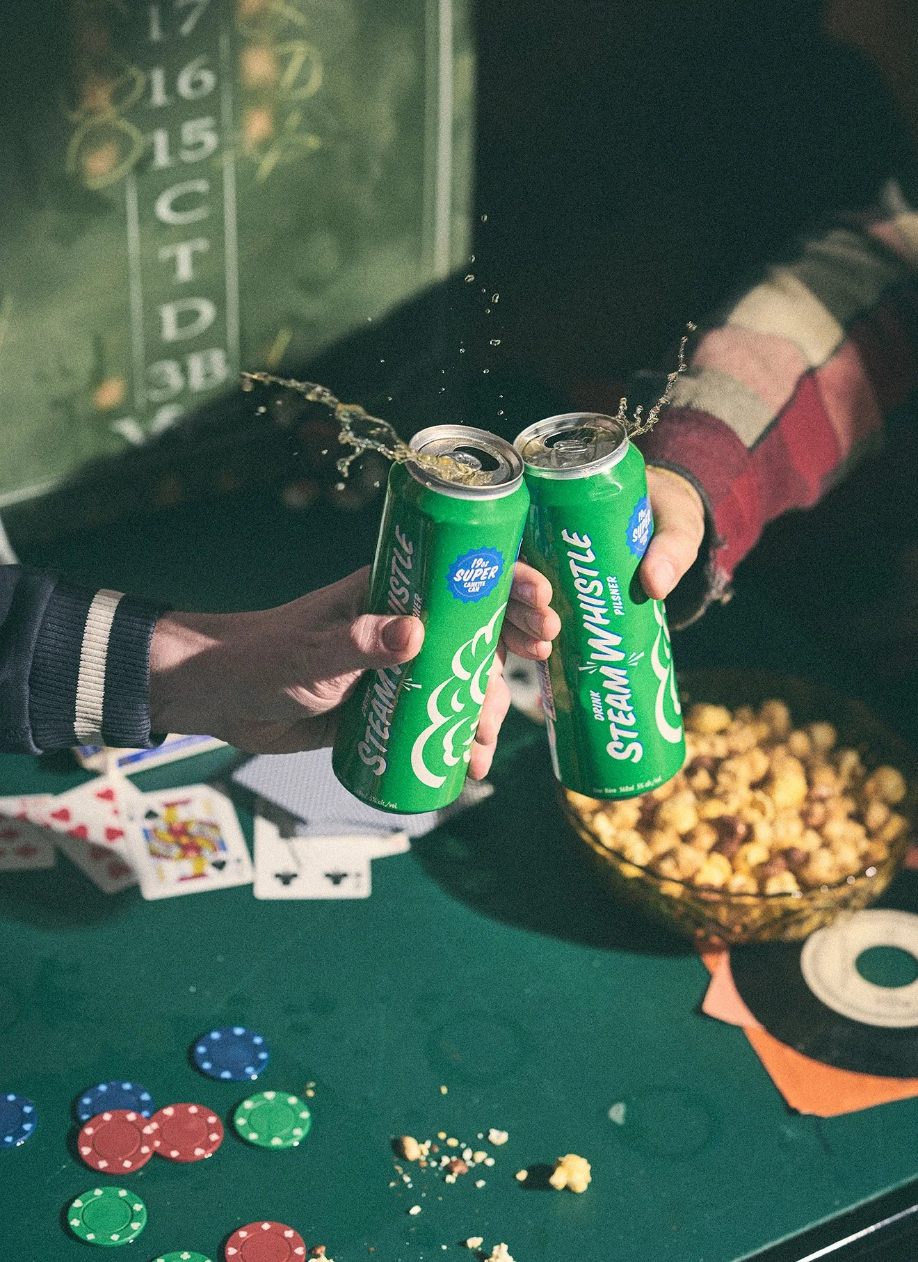 Two people holding cans of Steam Whistle clinking them together over a poker table, with poker chips, playing cards, and popcorn on the table. Photographed by Nick Ghattas, set stylist is Gumption Studio.