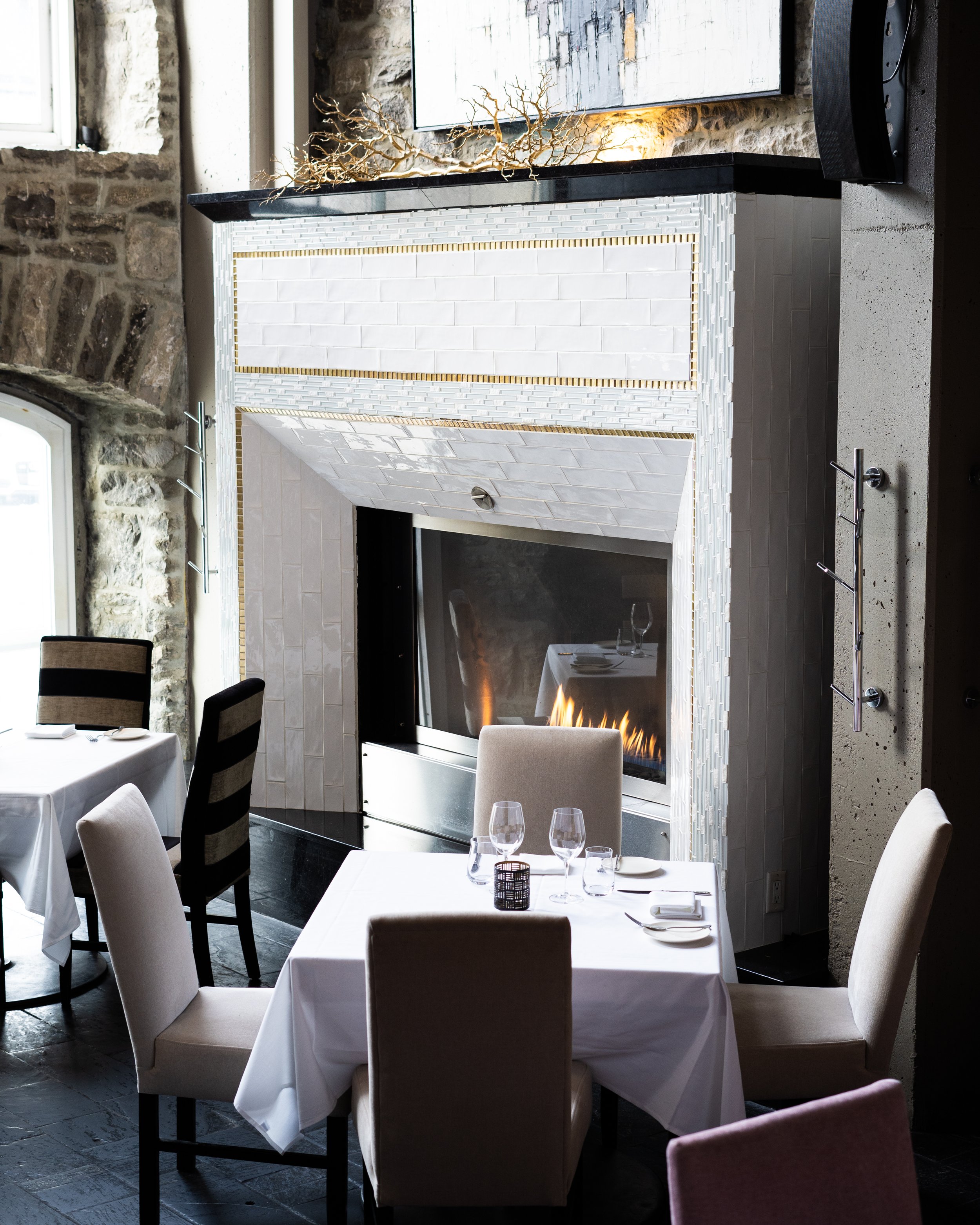 Restaurant e18hteen dining area with a fireplace, white tablecloths, wine glasses, and chairs, featuring a stone wall and modern decor. Photographed by Gumption Studio, Ottawa interior photographer. 