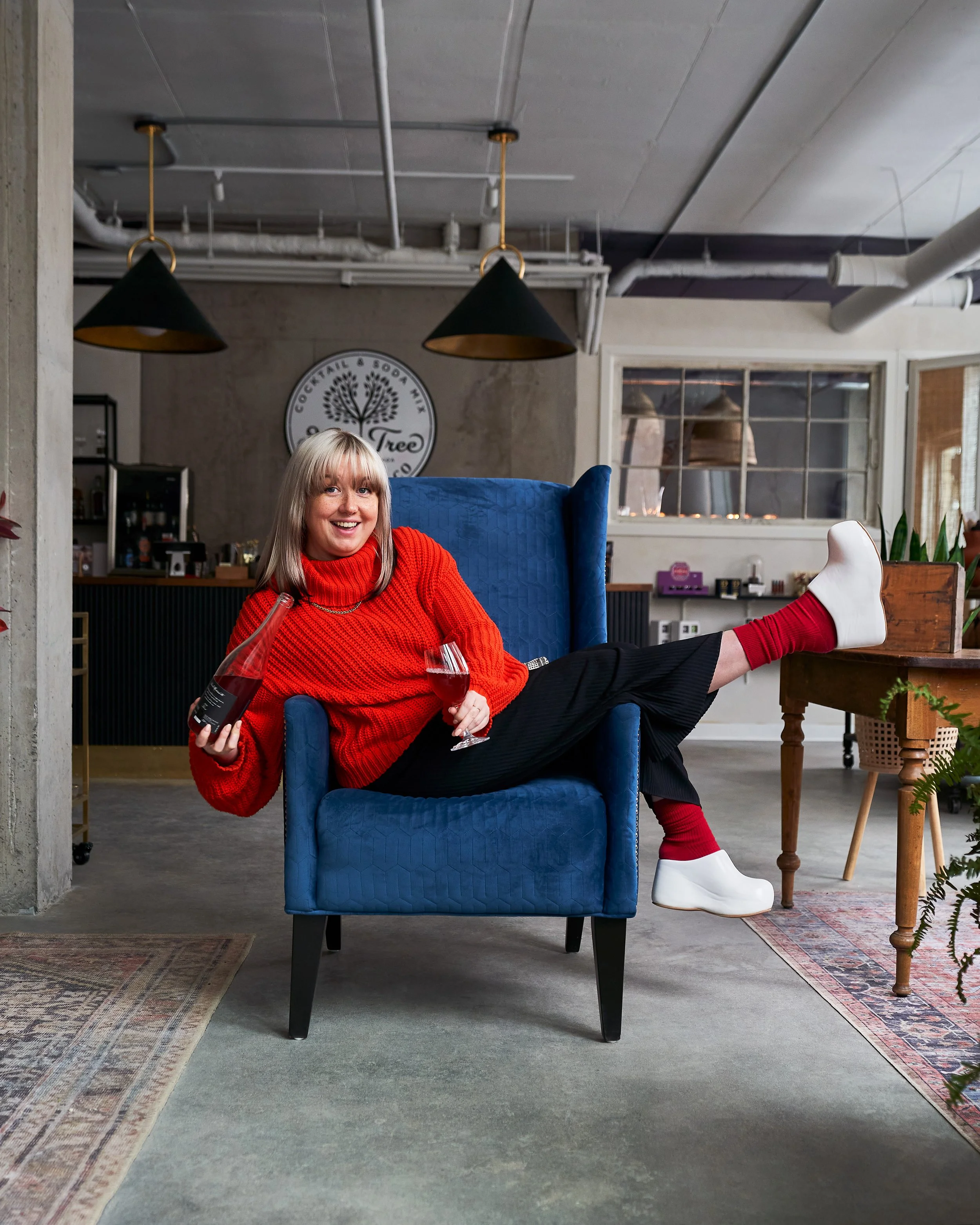 A woman with blonde hair smiling and sitting in a blue armchair inside a modern, industrial-style cafe or restaurant. She is wearing a red sweater, black pants, white boots, and red socks, holding a wine glass in one hand and a bottle in the other. T