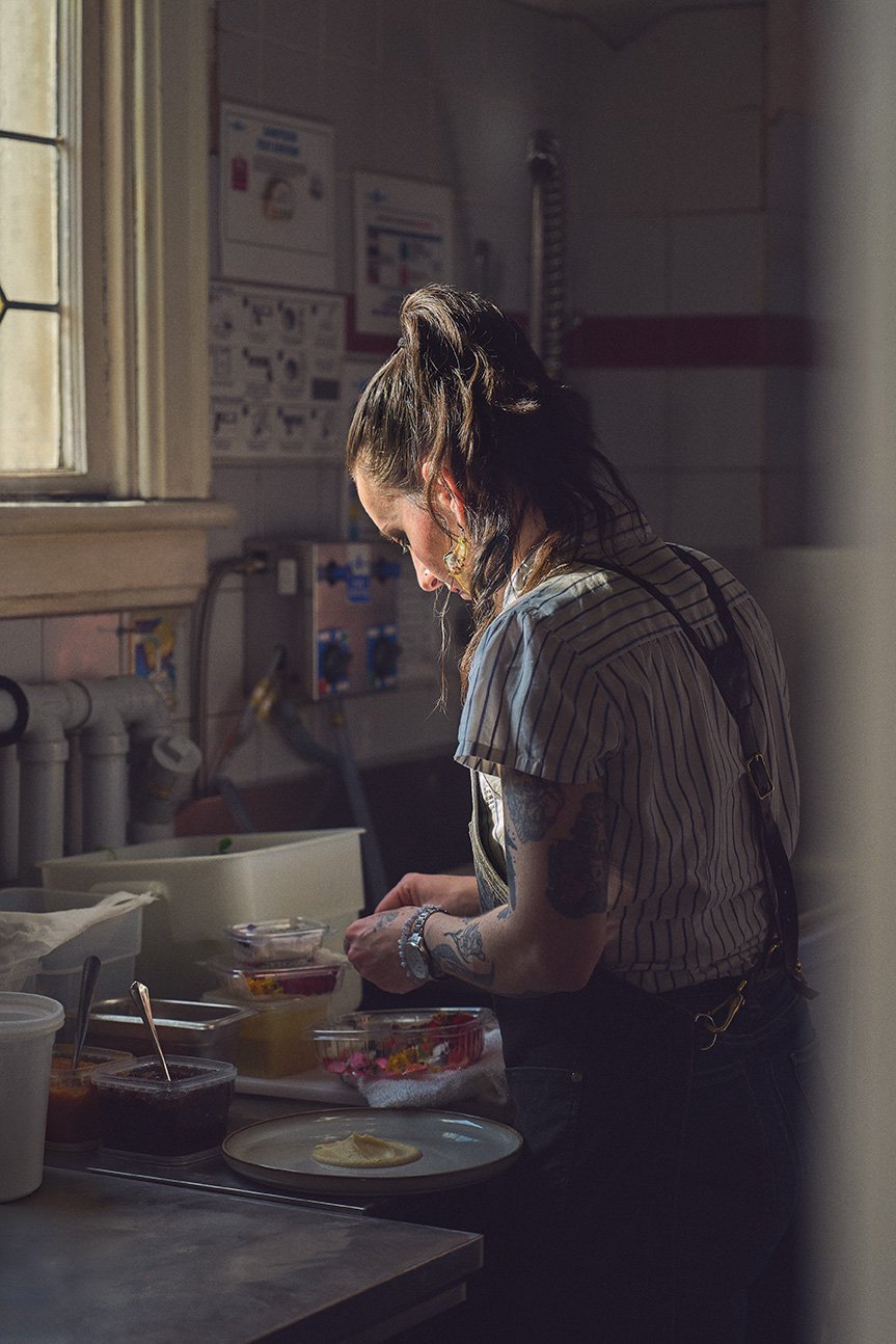 A woman with dark hair in a ponytail and tattoos on her arms is preparing food in a kitchen, illuminated by sunlight through a window.