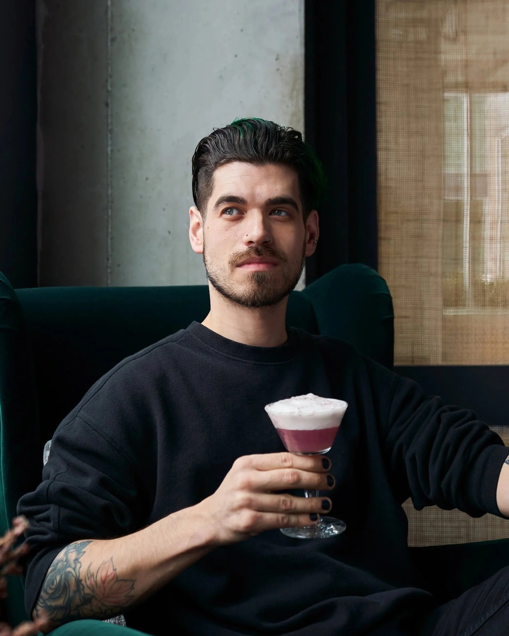 A young man with dark hair, a beard, and tattoos on his arm, sitting in a modern indoor setting, holding a cocktail glass with a layered pink and white drink, gazing thoughtfully to the side.