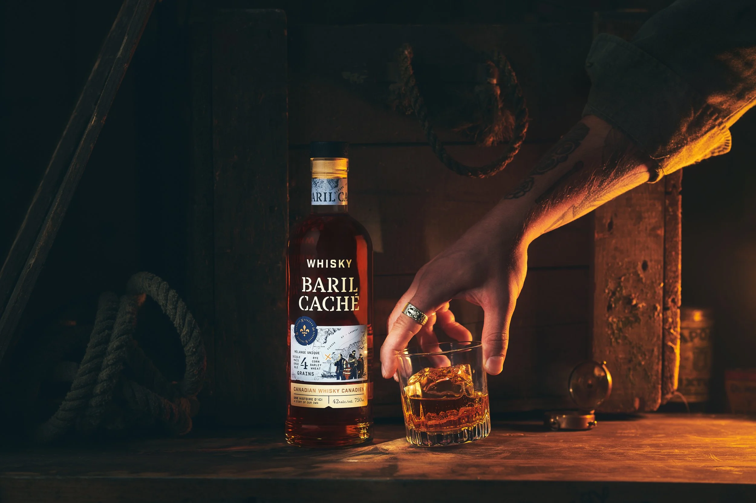 A hand with tattoos is pouring whiskey from a bottle labeled 'Baril Caché' into a glass. The scene is dimly lit with warm lighting on a wooden surface.  Photographed by Nick Ghattas, Ottawa food stylist is Gumption Studio.