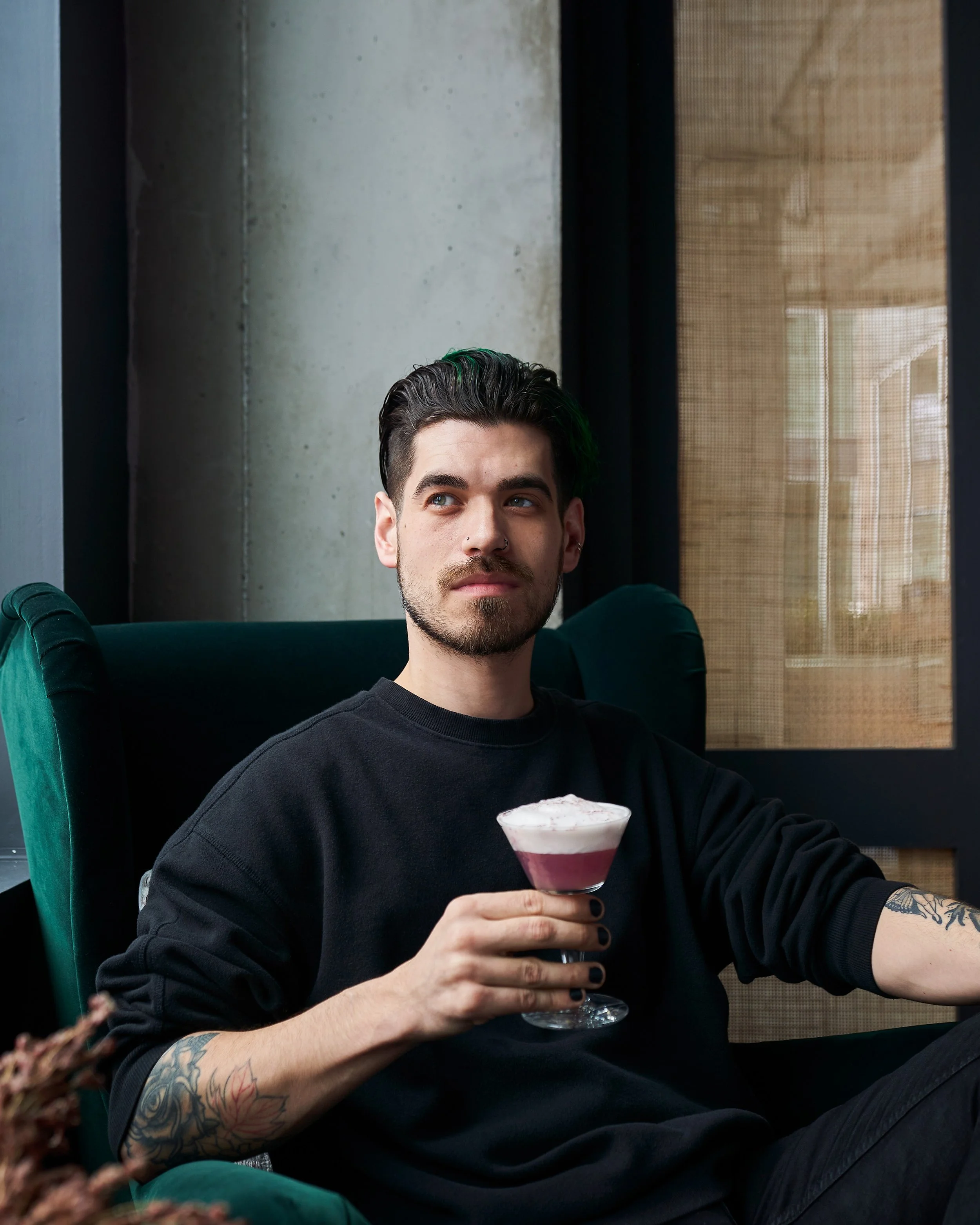 A young man with dark hair and a mustache, wearing a black long-sleeve shirt, is sitting indoors holding a glass of layered pink and white dessert. He has a tattoo on his right forearm and a nose piercing. Part of his other arm is visible in the fore