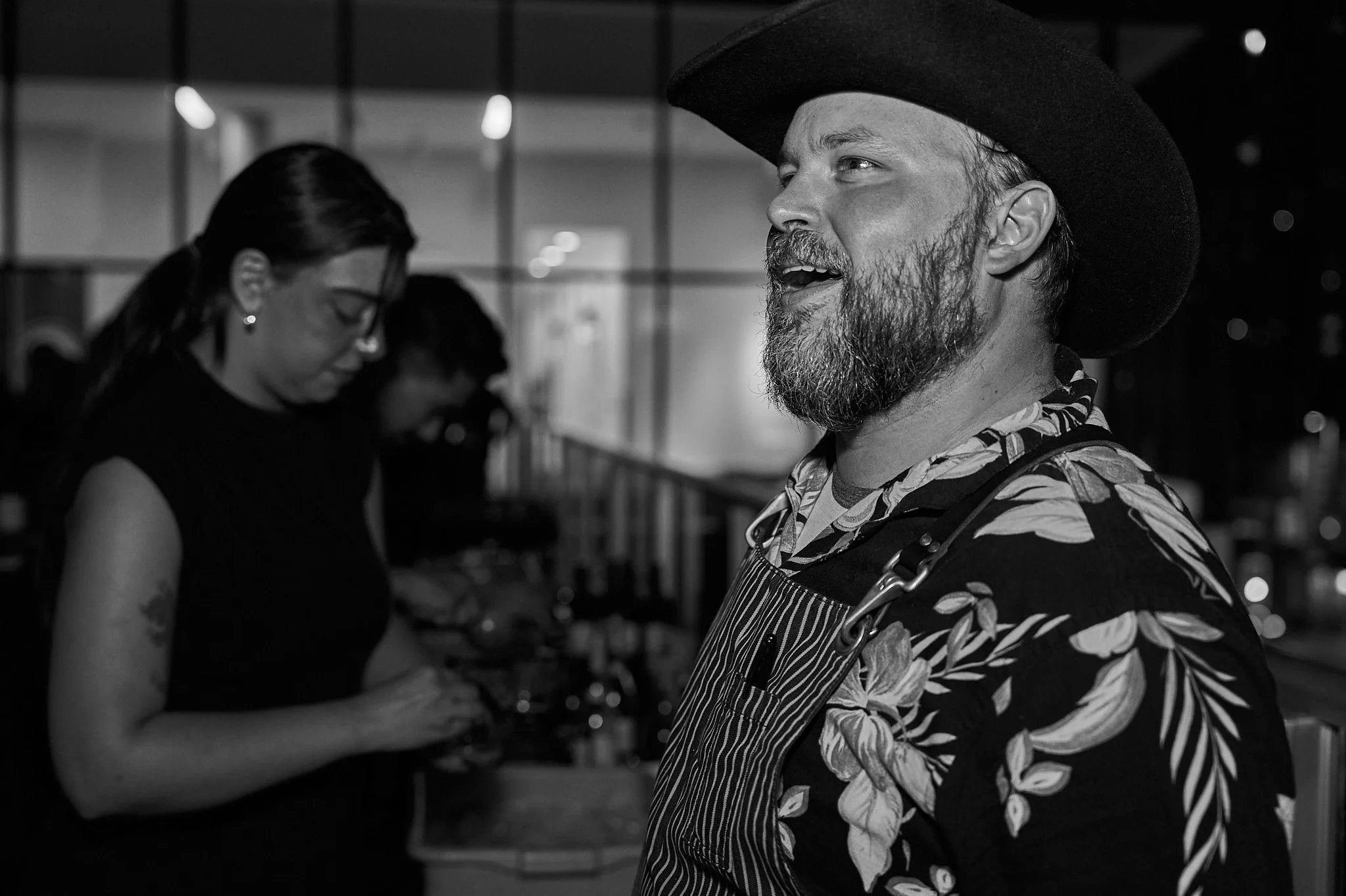 Chef Dave Godsoe with a beard wearing a large black hat and a patterned shirt at Harvest: A Feast of Fall with a woman in black attire in the background. Photographed by Gumption Studio, Ottawa event photographer.