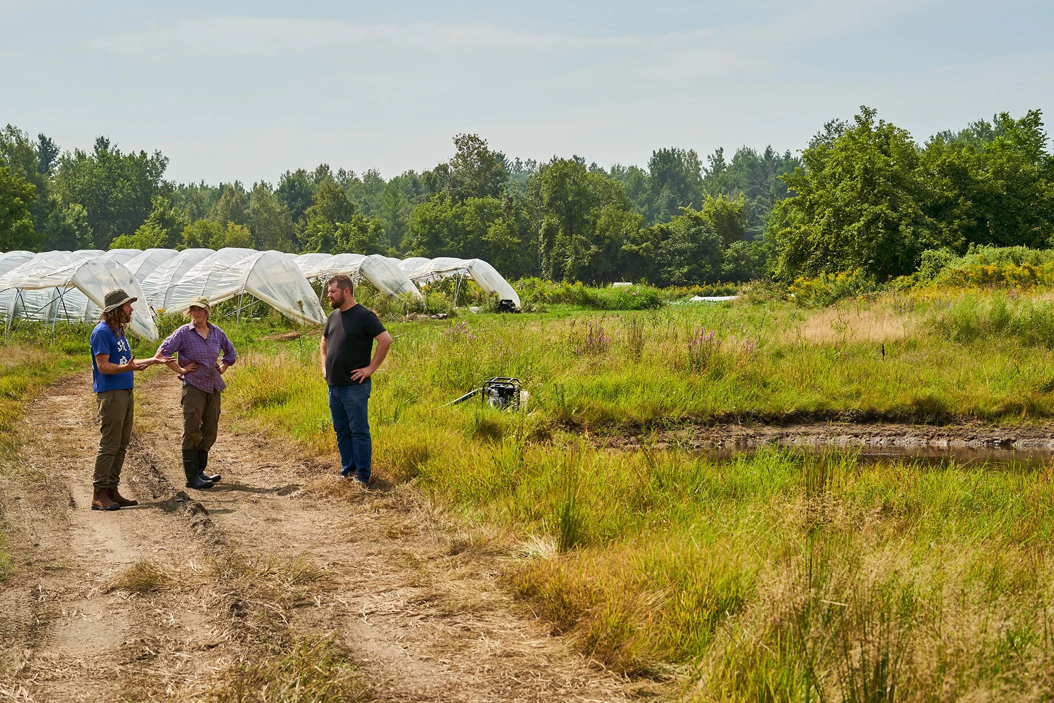 Justin Champagne-Lagarde at Heart City Farm, standing outdoors near a green field with a greenhouse in the background, engaging in conversation. Photographed for the Perch Cookbook by Gumption Studio, Ottawa Cookbook photographer.