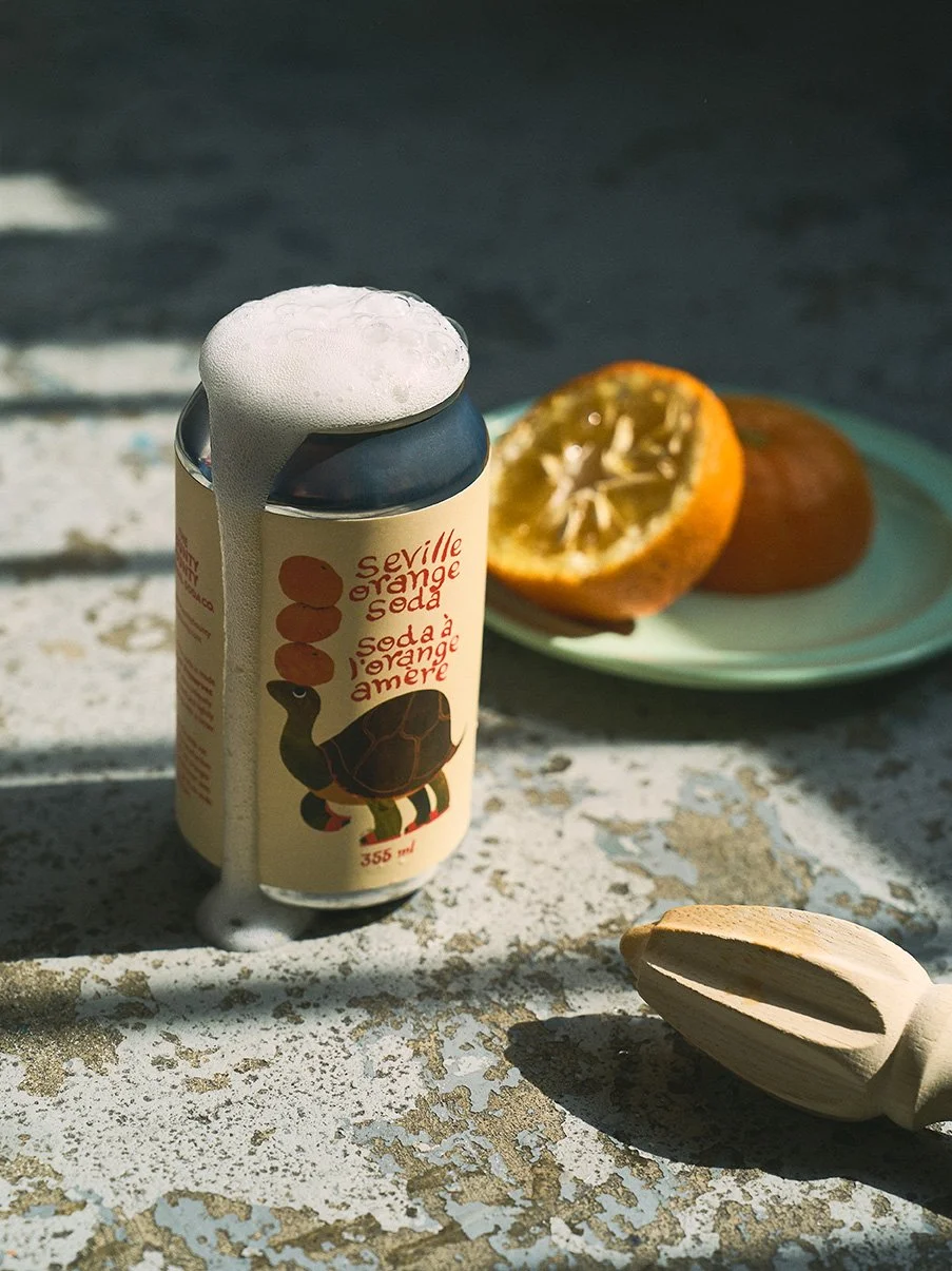 A can of Seville orange soda by Top Shelf Preserves with foam overflowing, placed on a weathered surface. Behind it, there is a green plate with a halved orange. To the right, there is a wooden citrus reamer. Product photography by Gumption Studio.