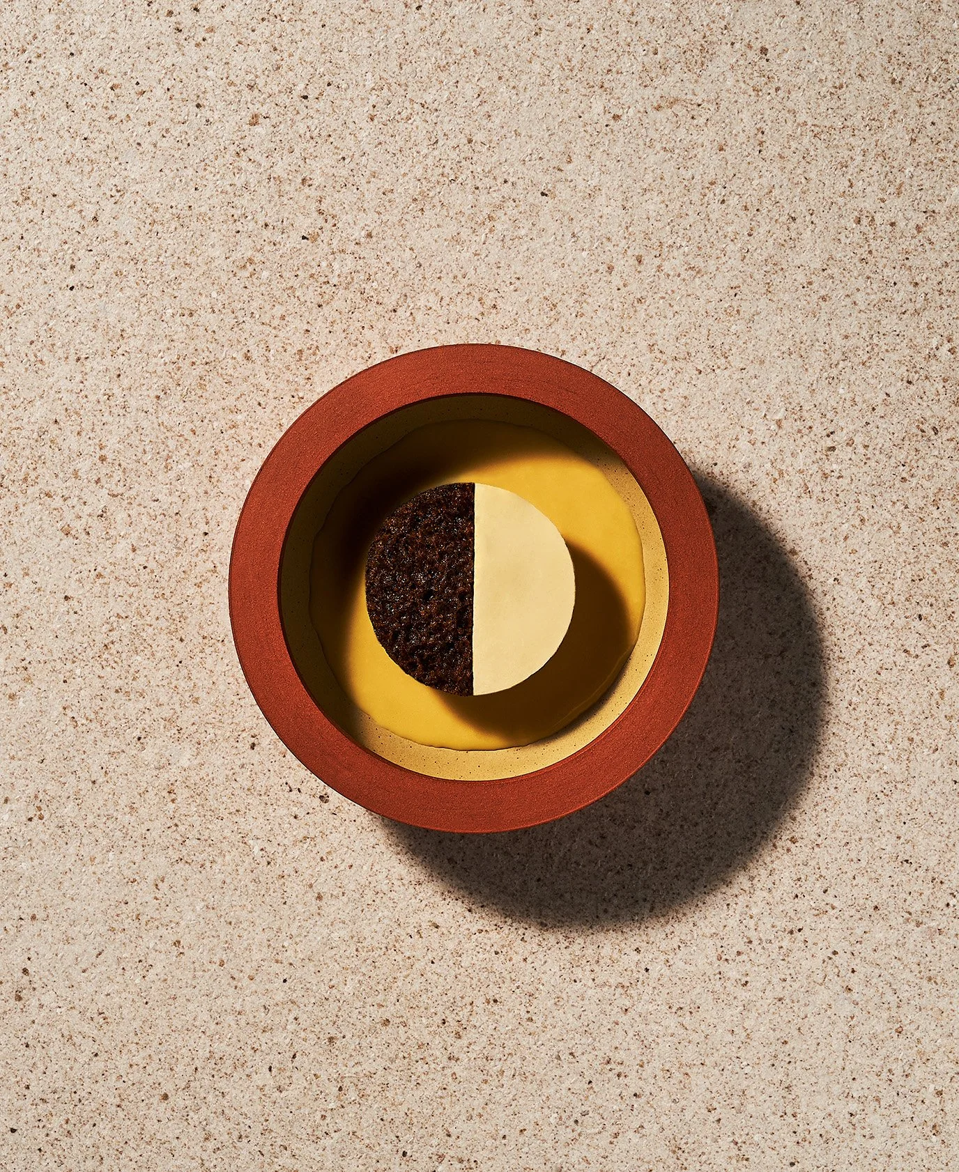 A Chaga dessert with half yellow and half dark brown surface inside a red container on a speckled beige countertop. Photographed for the Perch Cookbook by Gumption Studio, Ottawa Cookbook photographer.