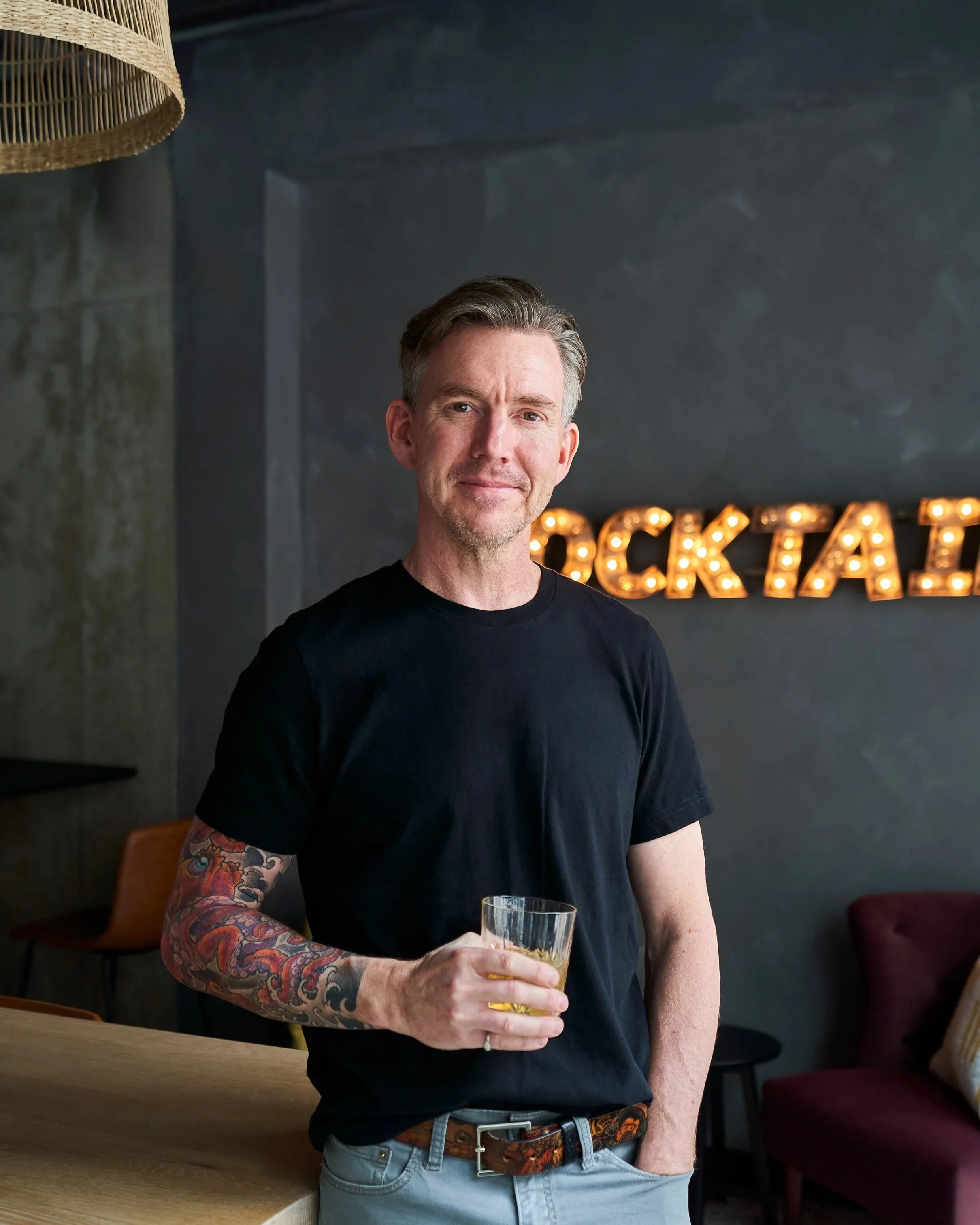 A man with gray hair and a tattoo sleeve on his left arm standing in a room with a dark wall, holding a glass of whiskey, smiling slightly, with illuminated marquee letters on the wall behind him.