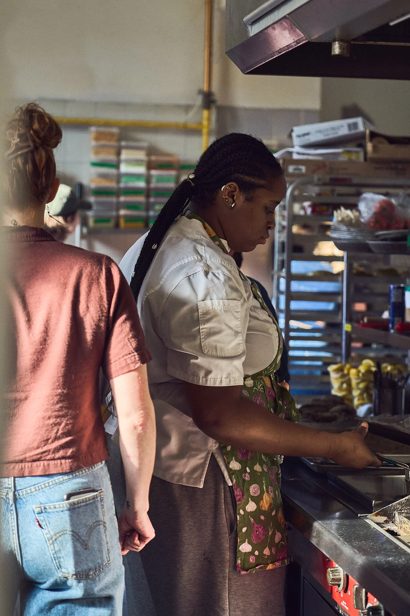 Two women, one with braided hair and earrings and the other with curly hair, are working in a commercial kitchen, preparing food at a stove with baked goods and food containers in the background.