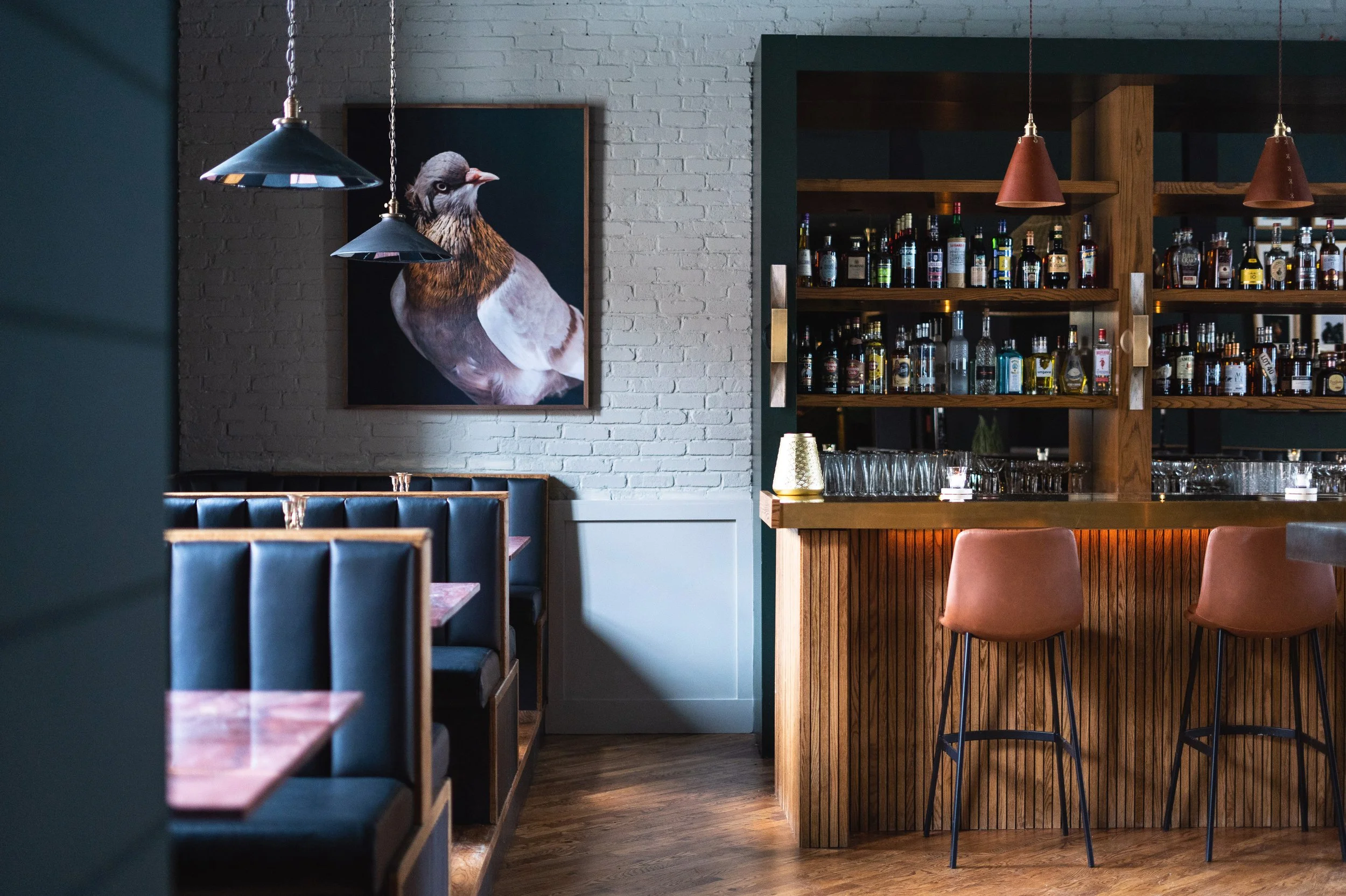 The Hyde interior with bottles of alcohol on wooden shelves, copper pendant lights, a framed bird portrait on a brick wall, and pink chairs around wooden tables. Photographed by Gumption Studio, Ottawa interior photographer.