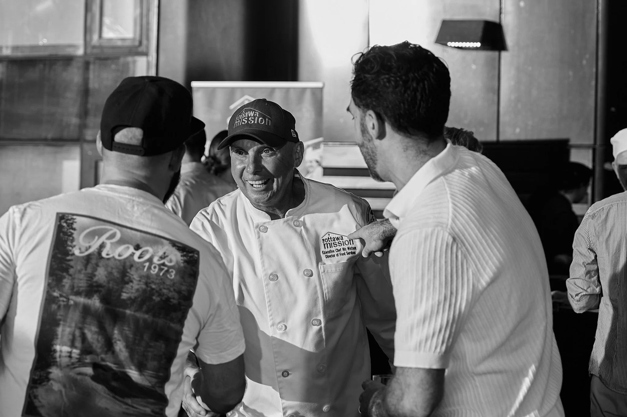 A group of people in conversation, with a chef wearing a uniform and a baseball cap that reads "Ottawa Mission," smiling. Photographed by Gumption Studio, Ottawa event photographer.