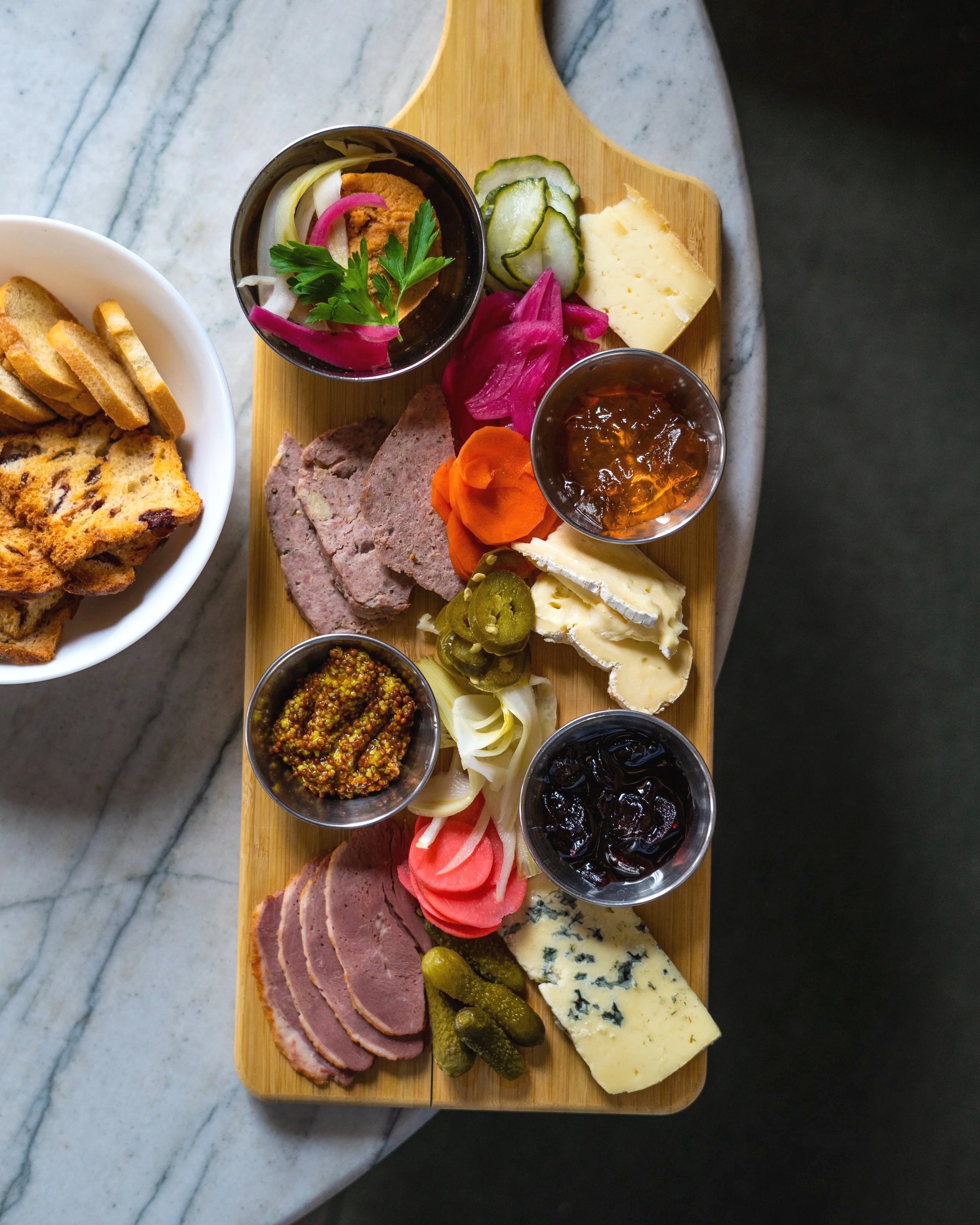 A cheese and charcuterie board from The Clarendon Tavern on a wooden platter featuring slices of meat, cheeses, pickles, on a marble surface. Photographed by Gumption Studio, Ottawa food photographer.