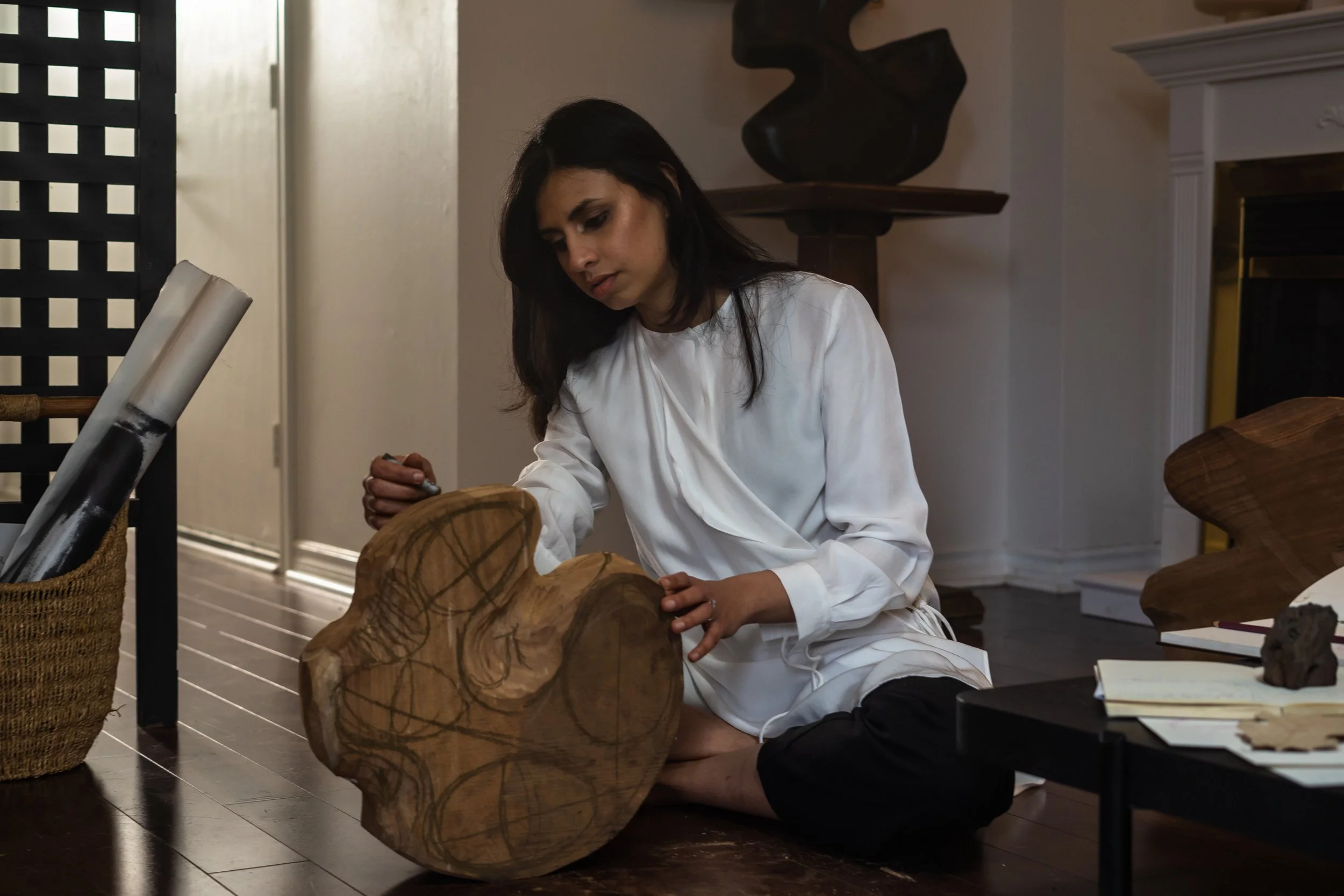 Woman with long dark hair, dressed in a white shirt, kneeling on the floor, working on a large wooden sculpture in an art studio.