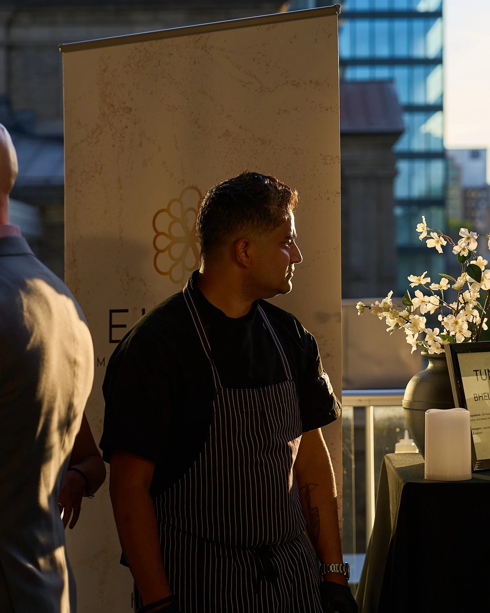 Chef Teegavarapu Sarath Mohan standing near a table with flowers, with sunlight casting shadows on his profile, at Harvest: A Feast of Fall during sunset. Photographed by Gumption Studio, Ottawa event photographer.