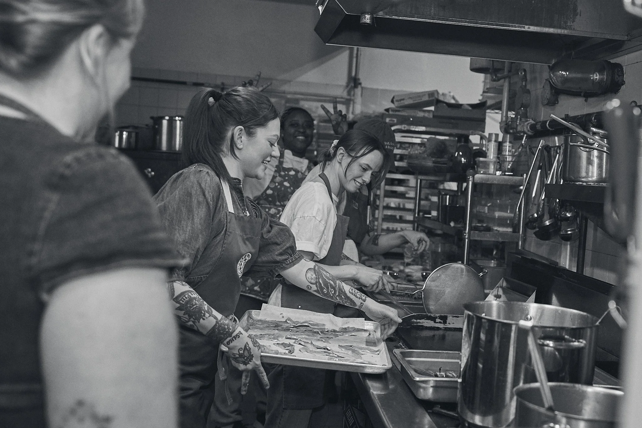 Four women working in a restaurant kitchen, preparing food at a stainless steel counter with cooking utensils and supplies around them. Photographed by Gumption Studio, Ottawa event photographer.