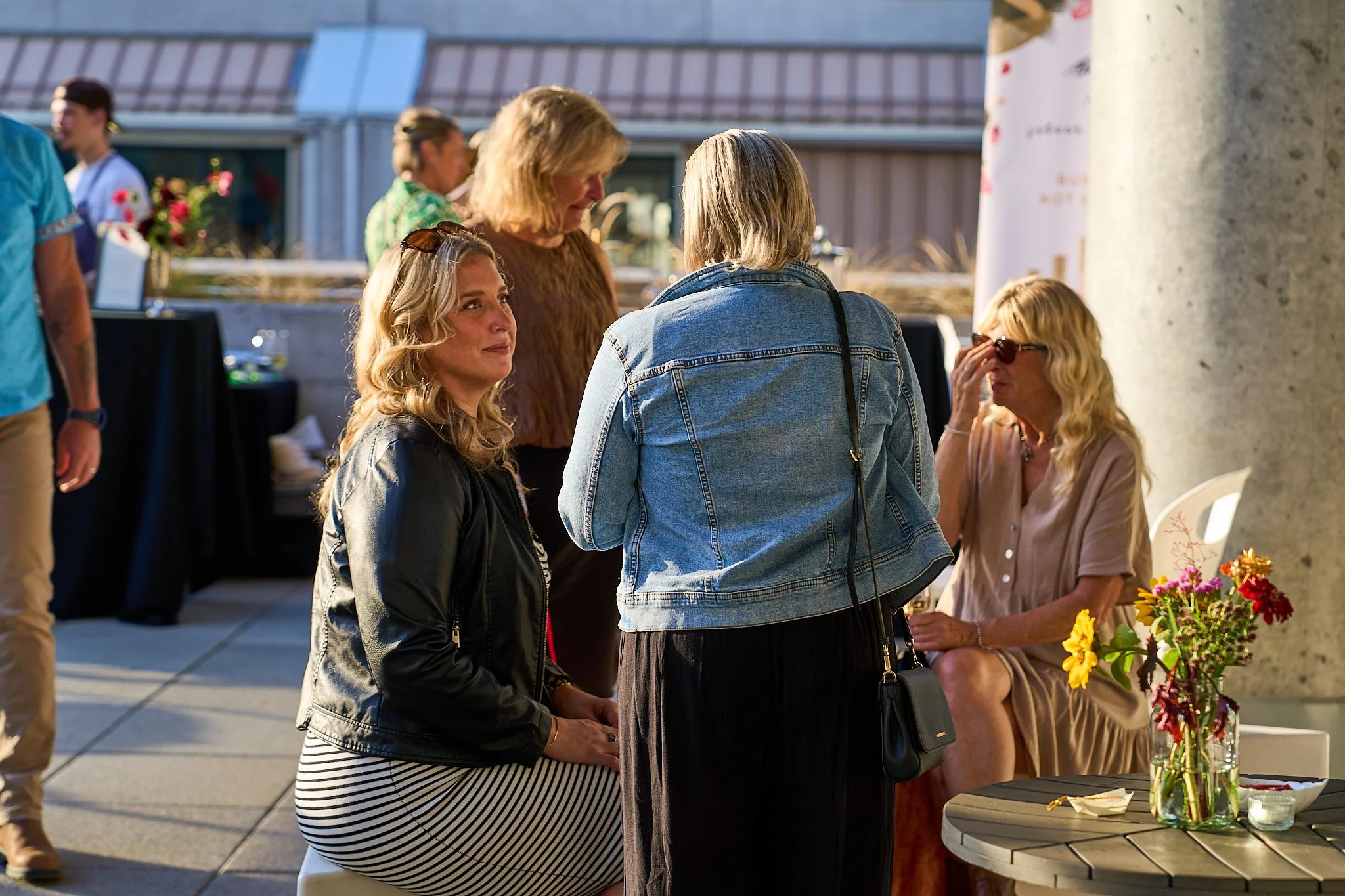 A group of women engaged in conversation at Harvest: A Feast of Fall with a table displaying flowers in the foreground and a building with a metal roof in the background. Photographed by Gumption Studio, Ottawa event photographer.