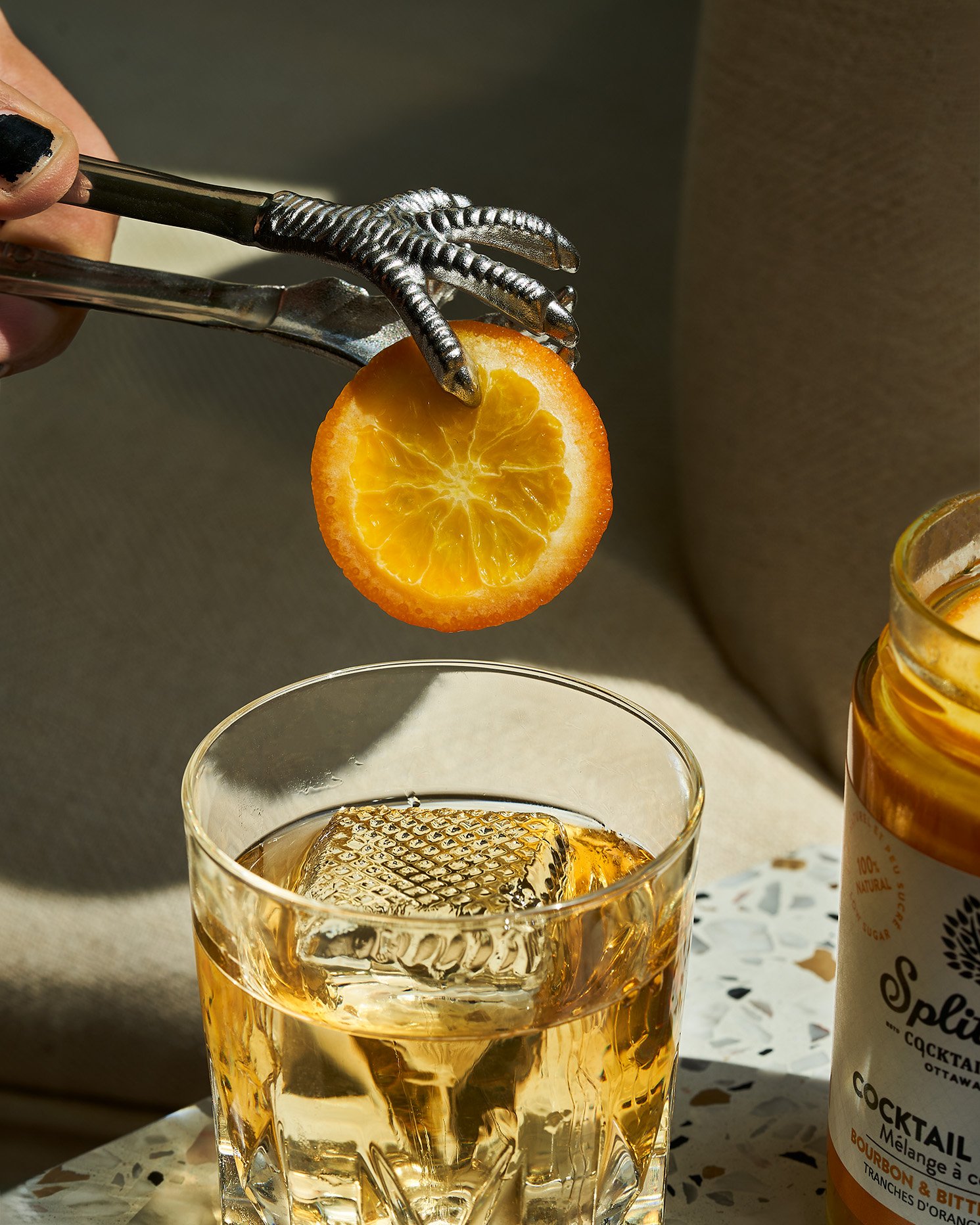 An old fashion cocktail with large cube and a orange slice being held above the glass by metallic tongs at Split Tree. Photographed by Gumption Studio, Ottawa cocktail photographer.