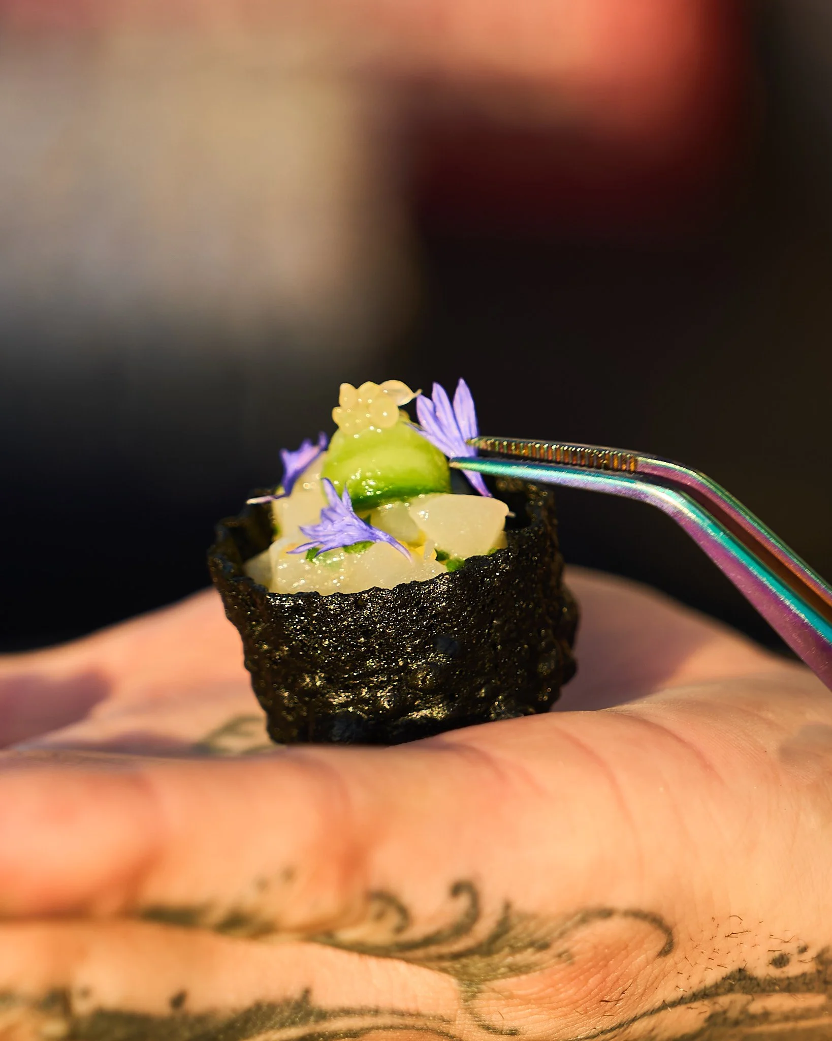 Close-up of a tiny sushi roll with seaweed wrap, topped with small purple flowers and green garnish, being held by a hand by Cameron McGihon. Photographed by Gumption Studio, Ottawa event photographer.