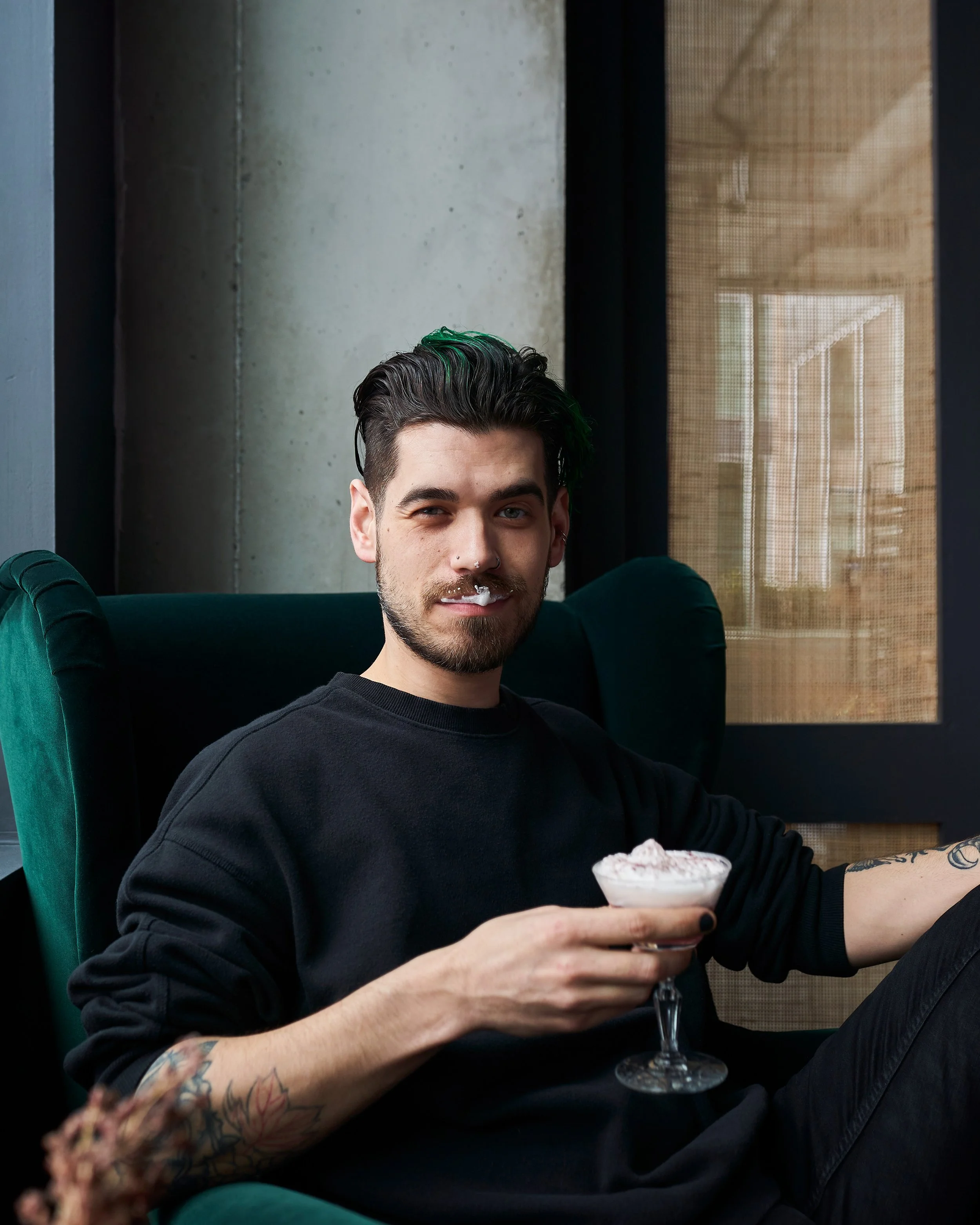 A young man with tattoos, dark hair with green highlights, and a nose piercing, sitting in a green velvet armchair, holding a cocktail glass topped with whipped cream, smiling, in a modern interior with concrete wall and a window with beige blinds.