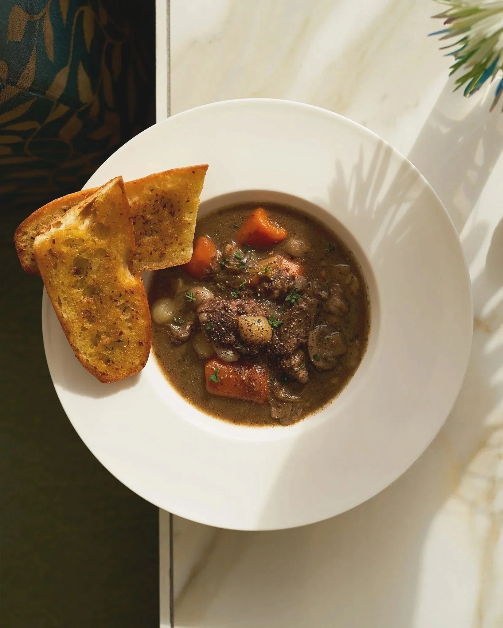 A bowl of beef stew with carrots, potatoes, and herbs, served with a slice of garlic bread on a white plate by Motel Chelsea. Photographed by Gumption Studio, Ottawa food photographer. 