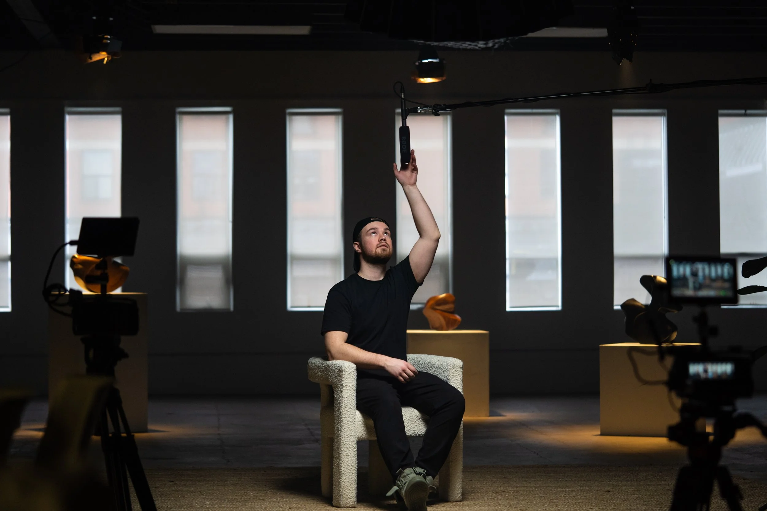 A man sitting on a textured chair in a dark room with tall windows in the background, reaching up towards a suspended microphone.