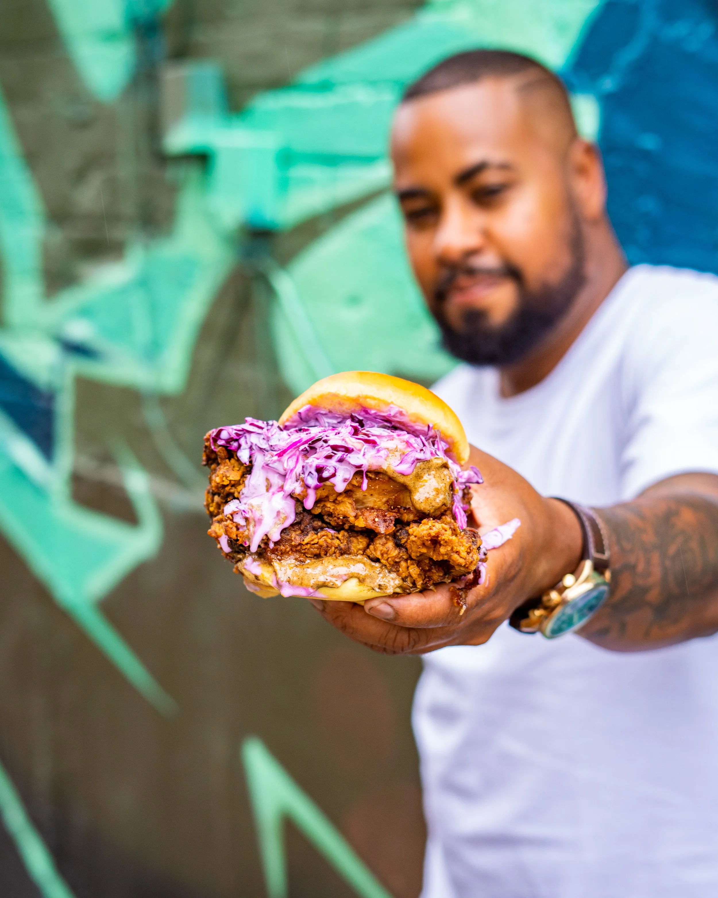 Jerome Robinson holding a large fried chicken burger with purple slaw by the Heartbreak Chef. Photographed by Gumption Studio, Toronto food photographer.