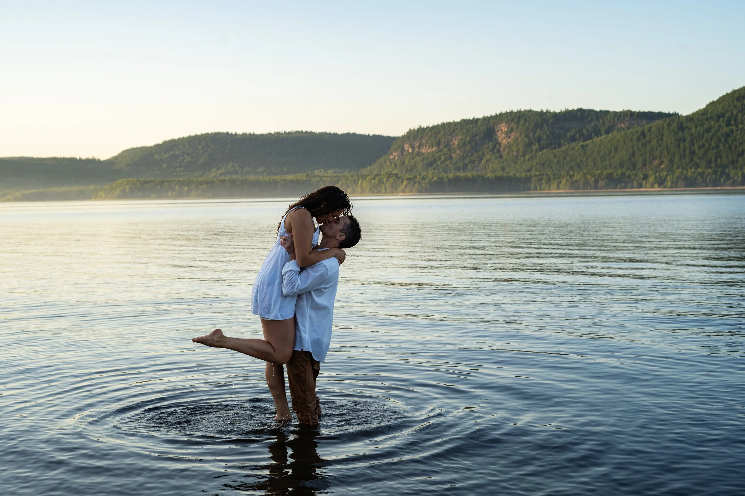 A couple standing in a lake, with the man kneeling in water holding the woman up as she kisses him, surrounded by a scenic landscape of trees and hills.