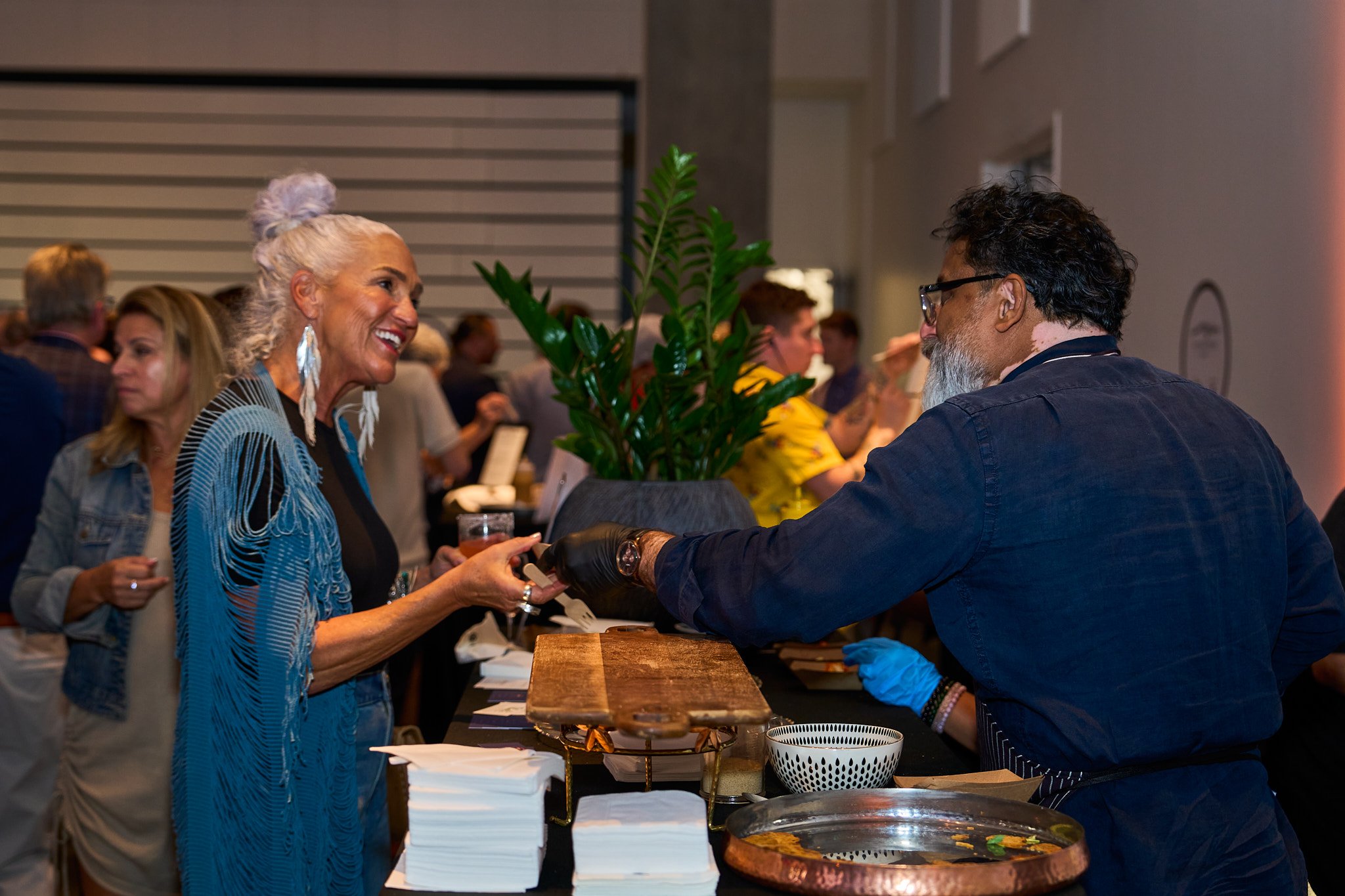 A woman with white hair in a bun, wearing a blue and black striped top, is smiling and receiving a drink from a man at a reception or event. The man is wearing glasses and a dark shirt, handing over a drink with gloves on. There are other people in t