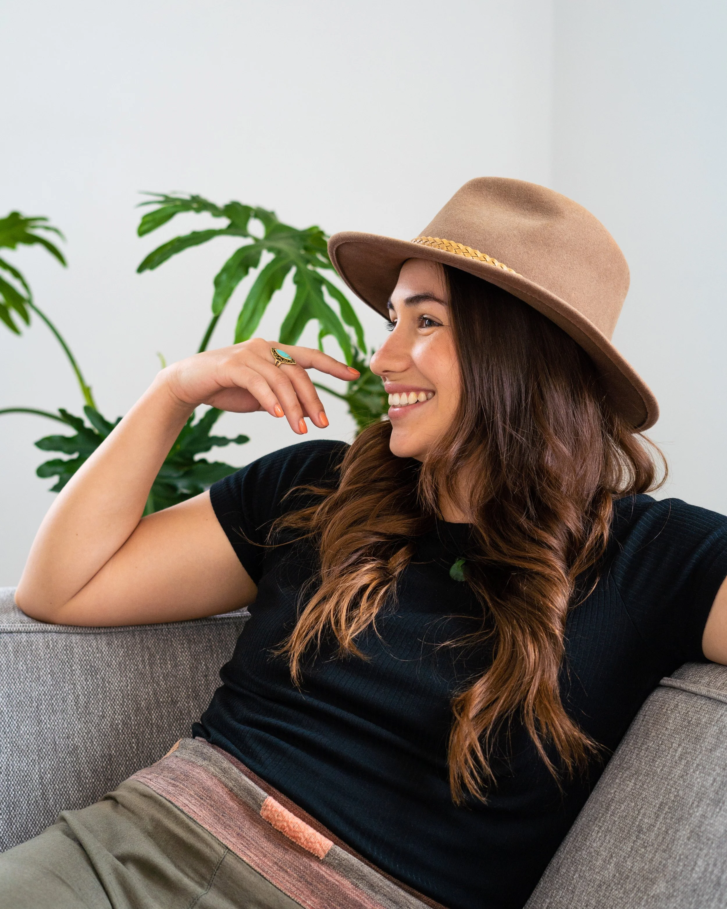 A woman with long brown hair smiling, wearing a tan wide-brimmed hat, a black top, and patterned pants, sitting on a gray couch with a green plant in the background.