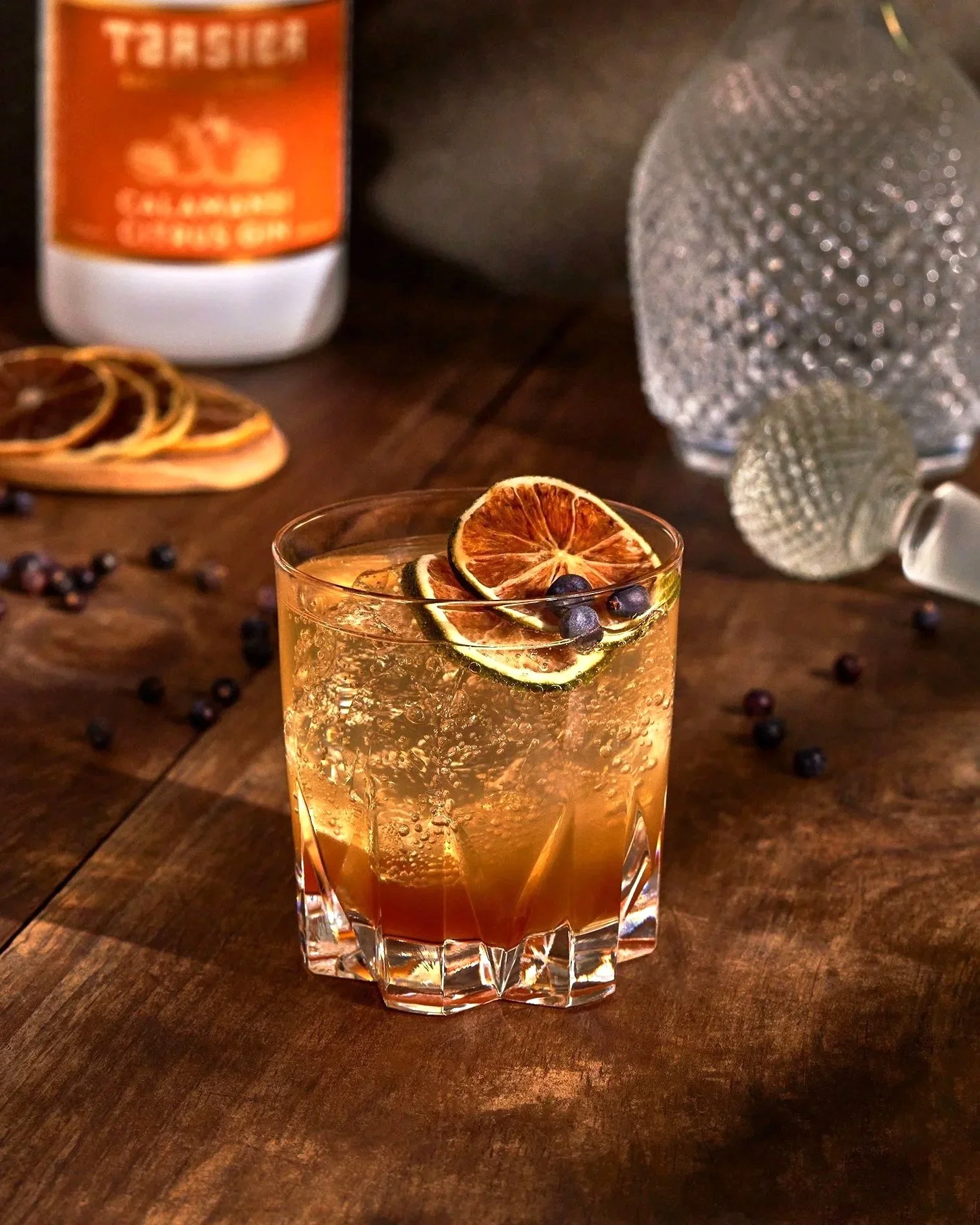 A cocktail  by Split Tree garnished with dried citrus slices and blueberries on a wooden table with scattered juniper. Photographed by Gumption Studio, Ottawa commercial photographer.