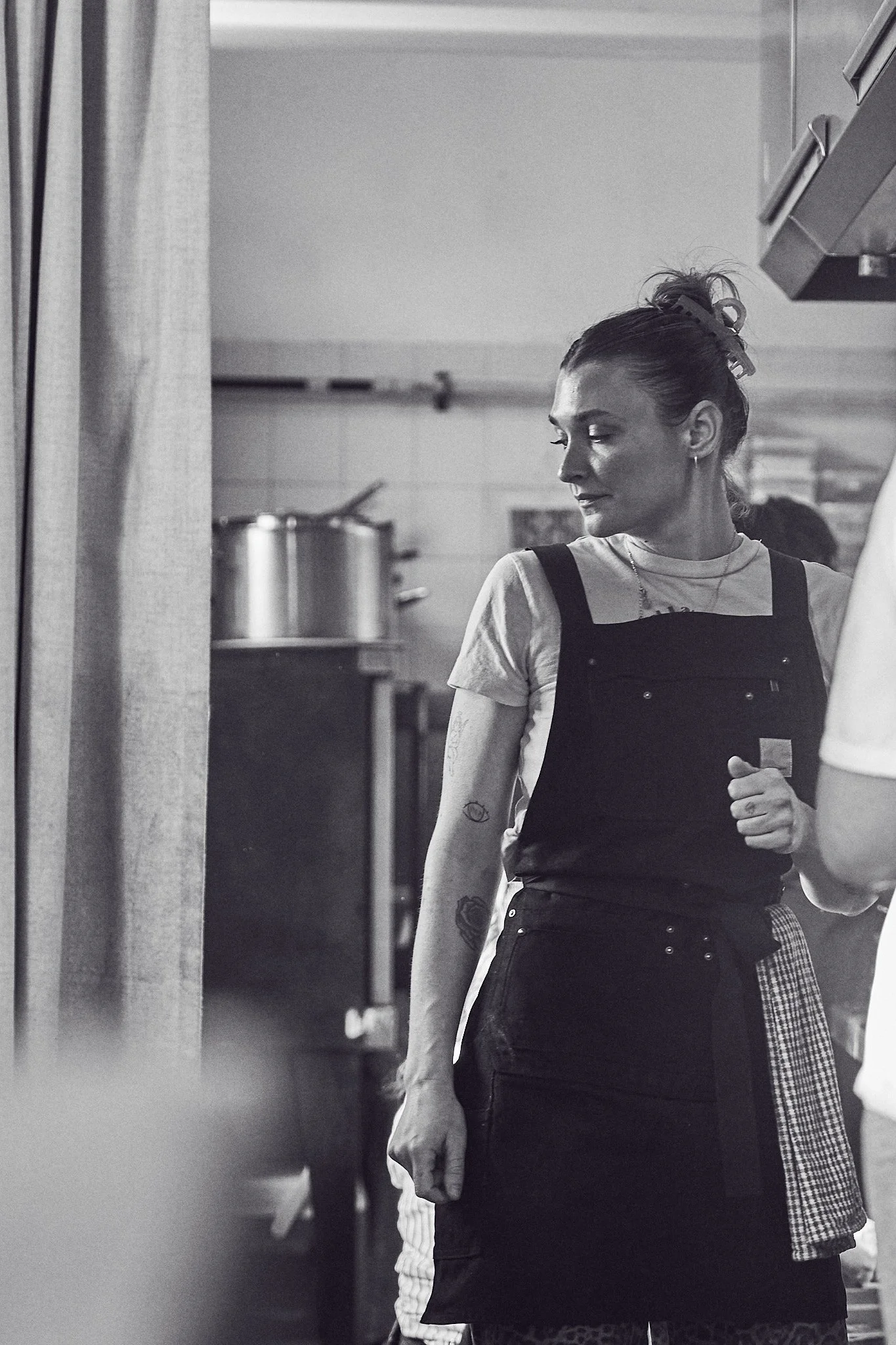 A woman with tattoos on her arm, wearing an apron, is looking at herself in a mirror in a kitchen.