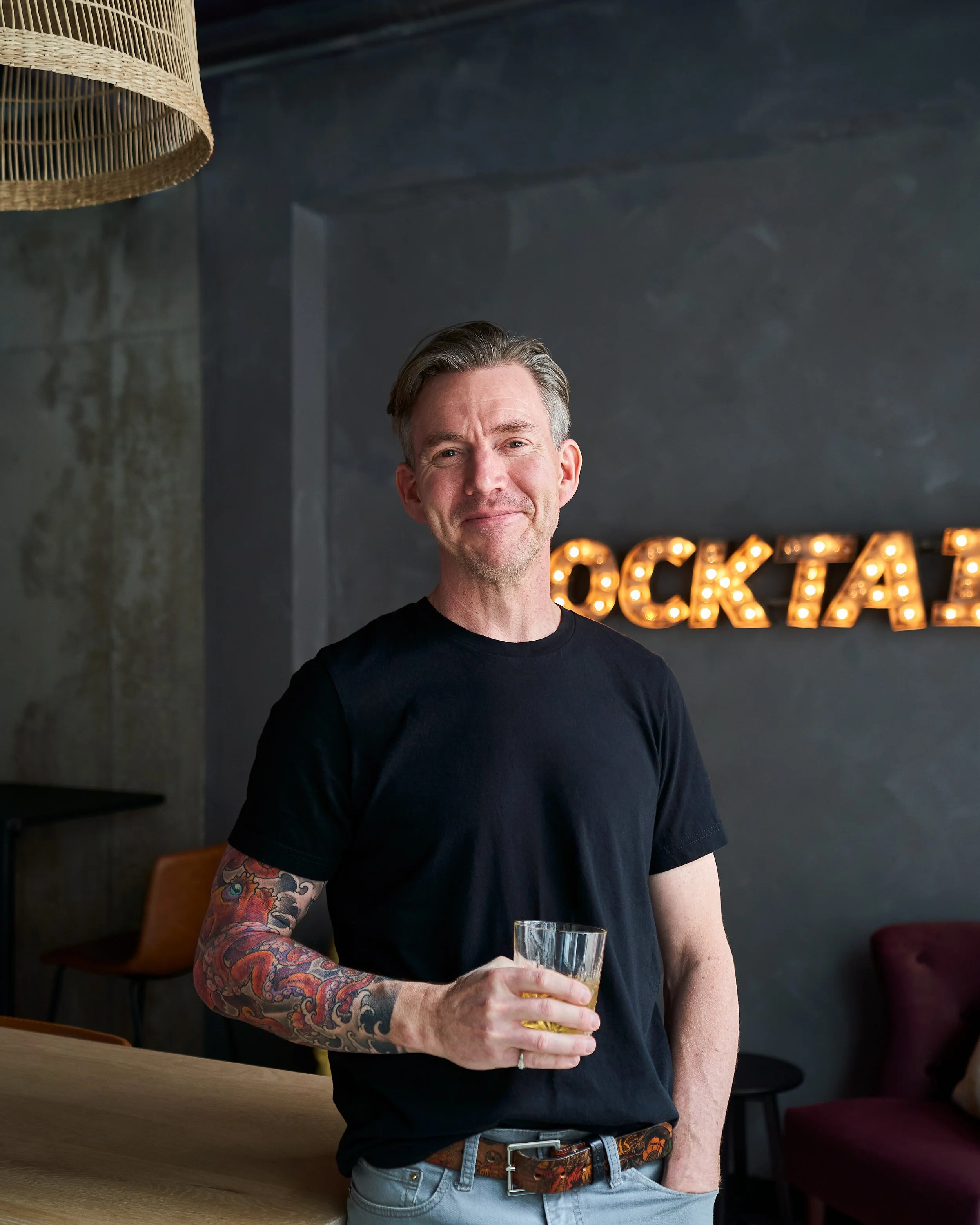 A man with gray hair, wearing a black t-shirt and gray jeans, is holding a glass of beer in a bar or restaurant. He has a tattooed sleeve on his right arm and is smiling. There is a decorative lit sign in the background that spells out 'COCKTAIL', al