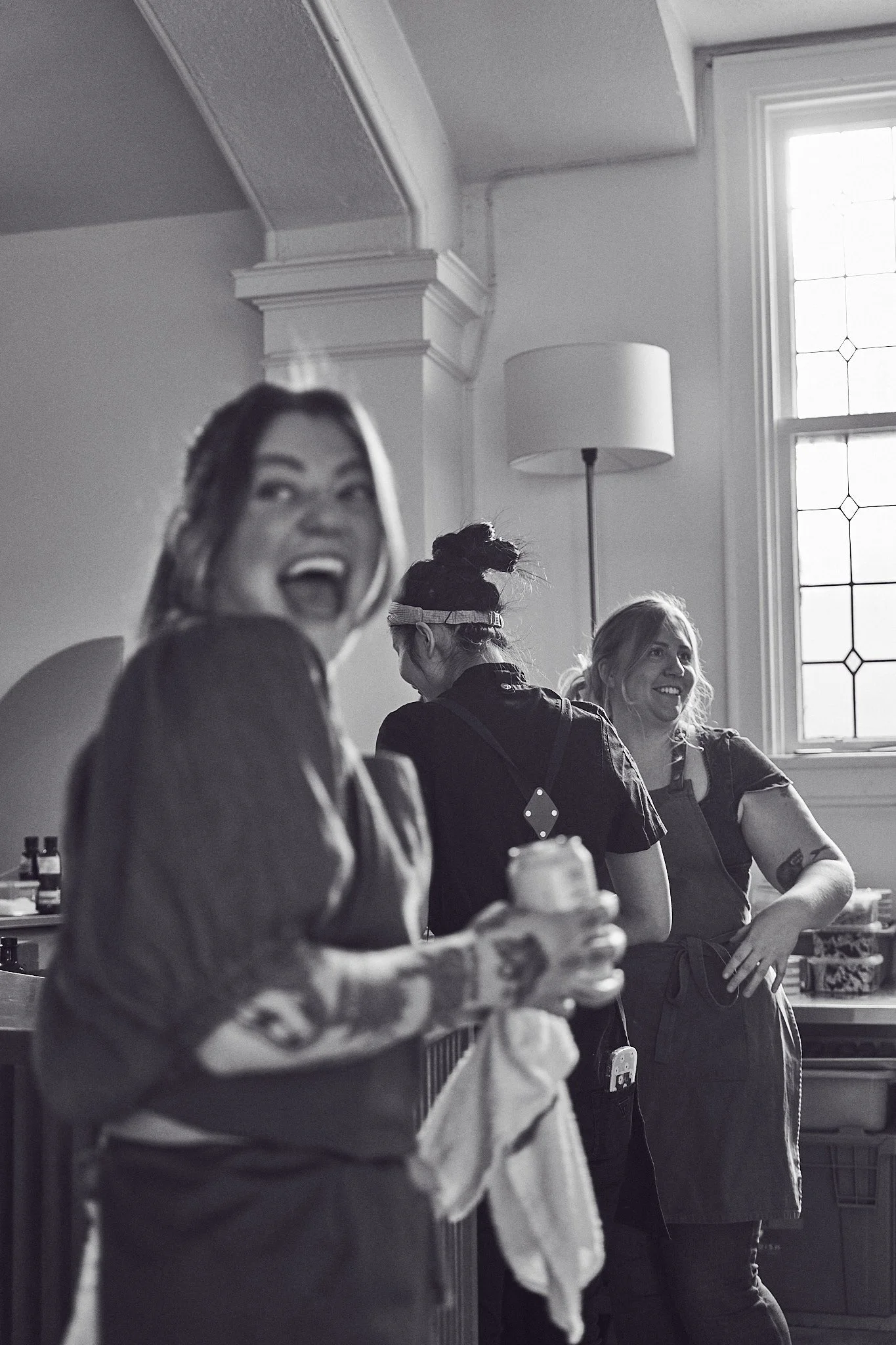 People gathered in a room near a window with sunlight, including a woman in the foreground smiling and looking to the side, another woman with her back turned, and a third woman smiling and looking to the side, all engaged in conversation or activity