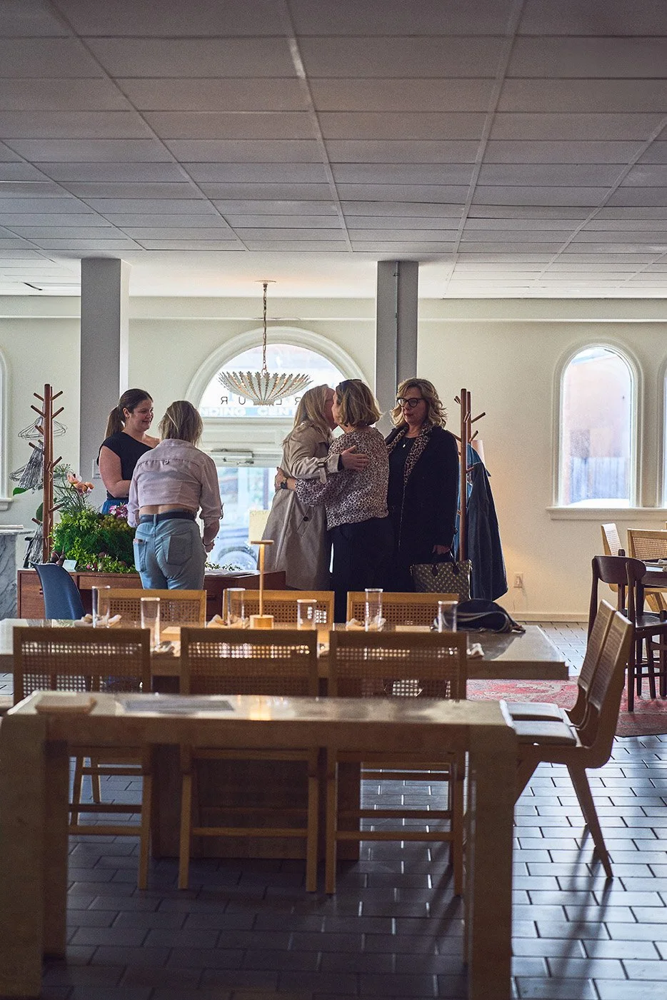 People gathered in a Parlour Restaurant for Girl Dinner, some greeting each other with hugs, with tables and chairs in the foreground and large arched windows in the background. Photographed by Gumption Studio, Ottawa event photographer.