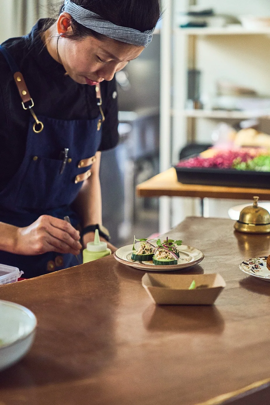 Chef Nora Jo wearing a navy apron and a headband, decorating small cucumber appetizer slices on a white plate in a kitchen. Photographed by Gumption Studio, Ottawa event photographer.