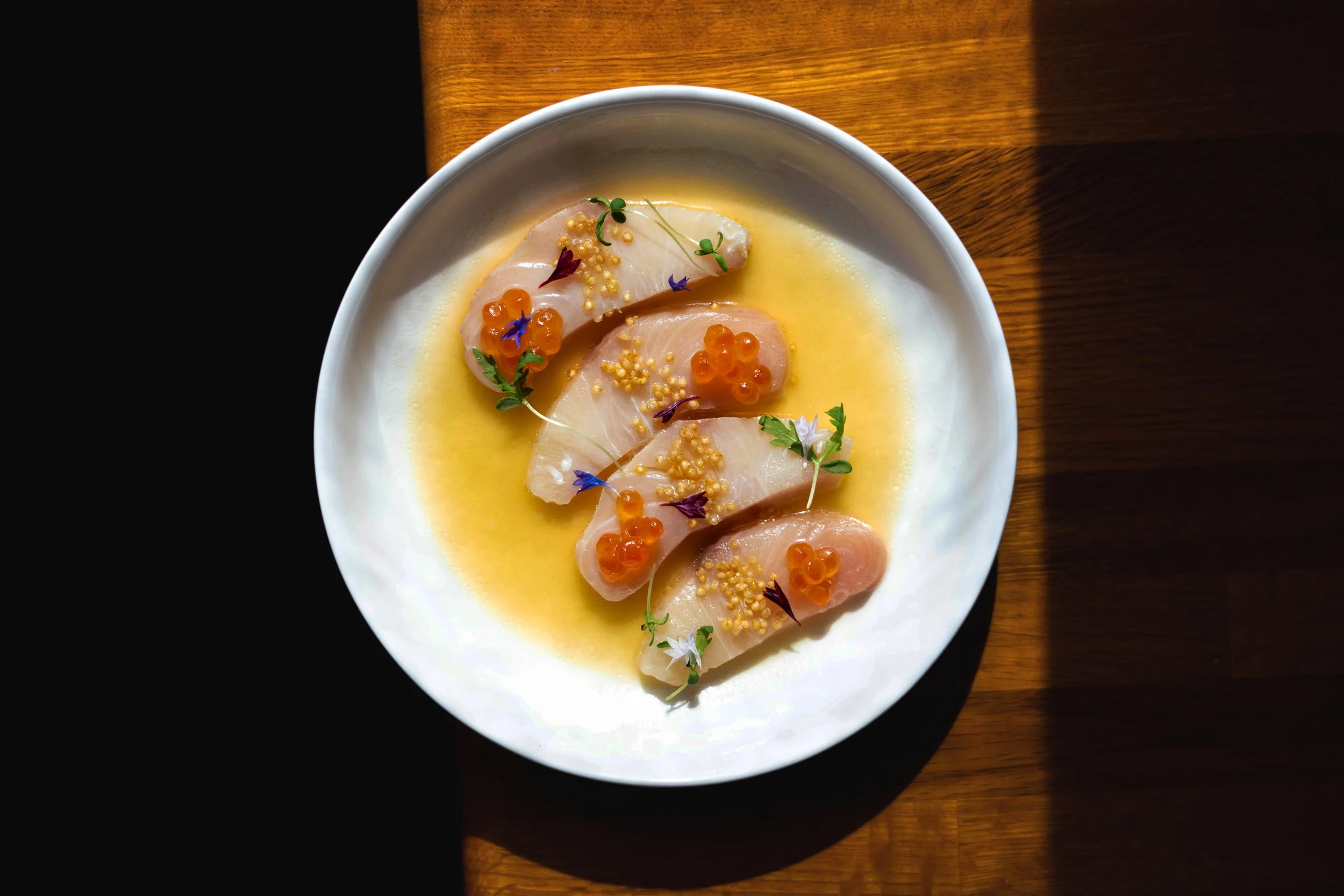 Tuna ceviche by The Soca Kitcen garnished with orange fish roe, microgreens, and edible flowers served on a yellow sauce in a white bowl on a wooden table. Photographed by Gumption Studio, Ottawa food photographer. 