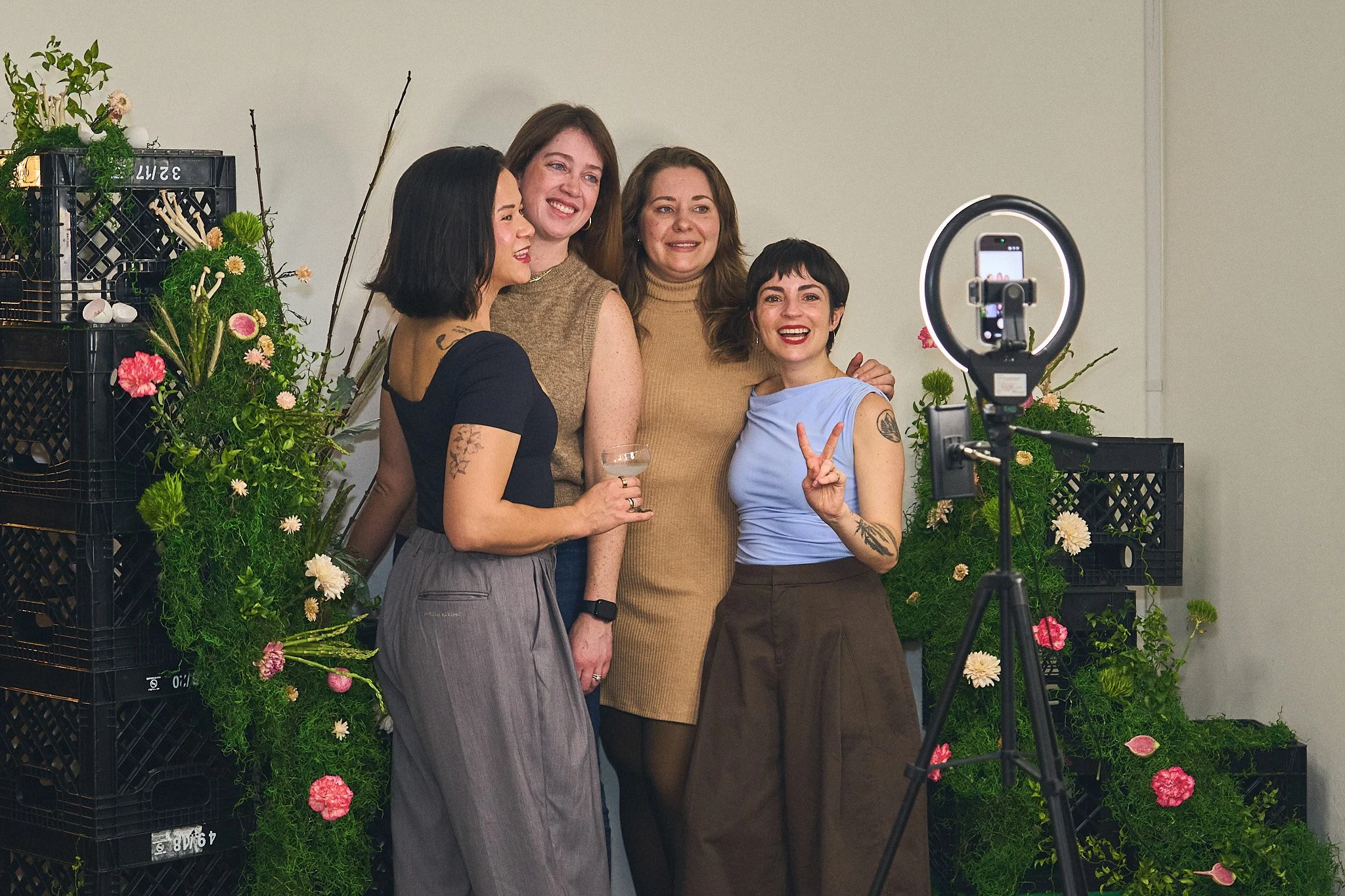 Five women taking a group selfie with a smartphone ring light, standing in front of a backdrop of flowers and greenery. Photographed by Gumption Studio, Ottawa event photographer.