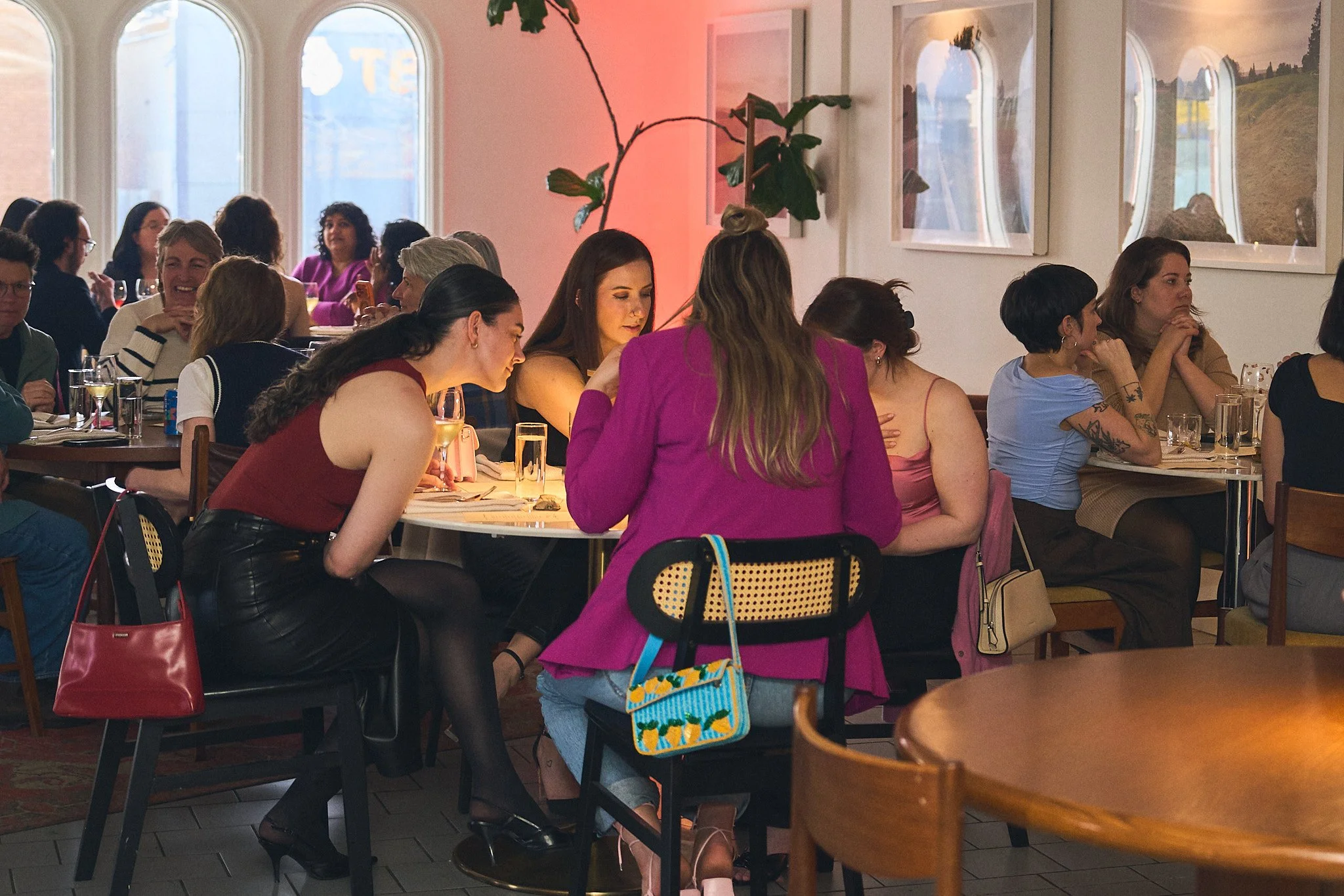 A group of people sitting at tables at Girl Dinner, with large windows and artwork on the walls. Some are engaged in conversation, while others are looking at their phones or eating. Photographed by Gumption Studio, Ottawa event photographer.