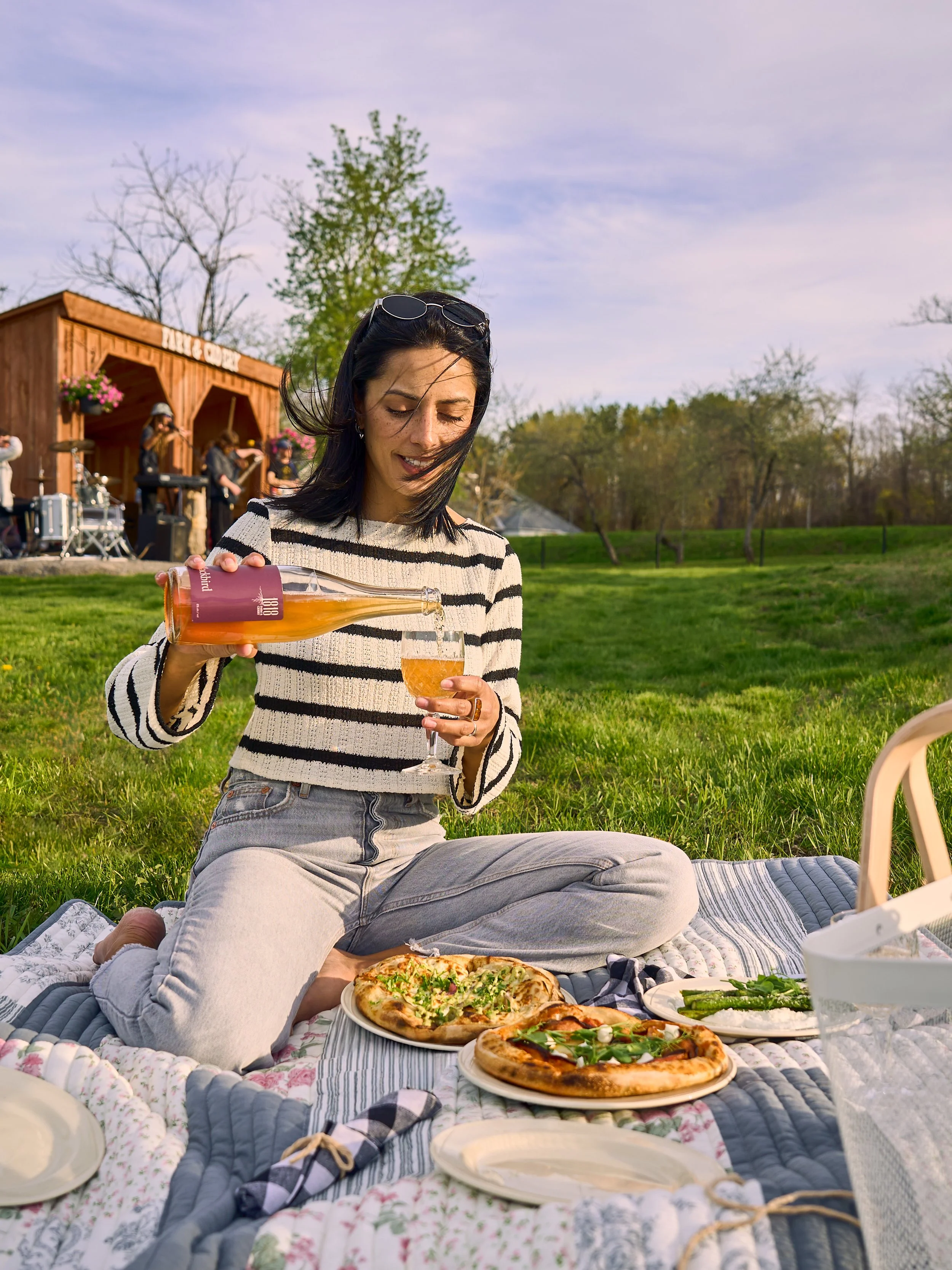 A woman sitting on a picnic blanket at 1818 Farm and Cidery pouring cider into a glass, with pizza and food spread on the blanket. Photo by Ottawa Photographer Sarah Farmer of Gumption Studio.