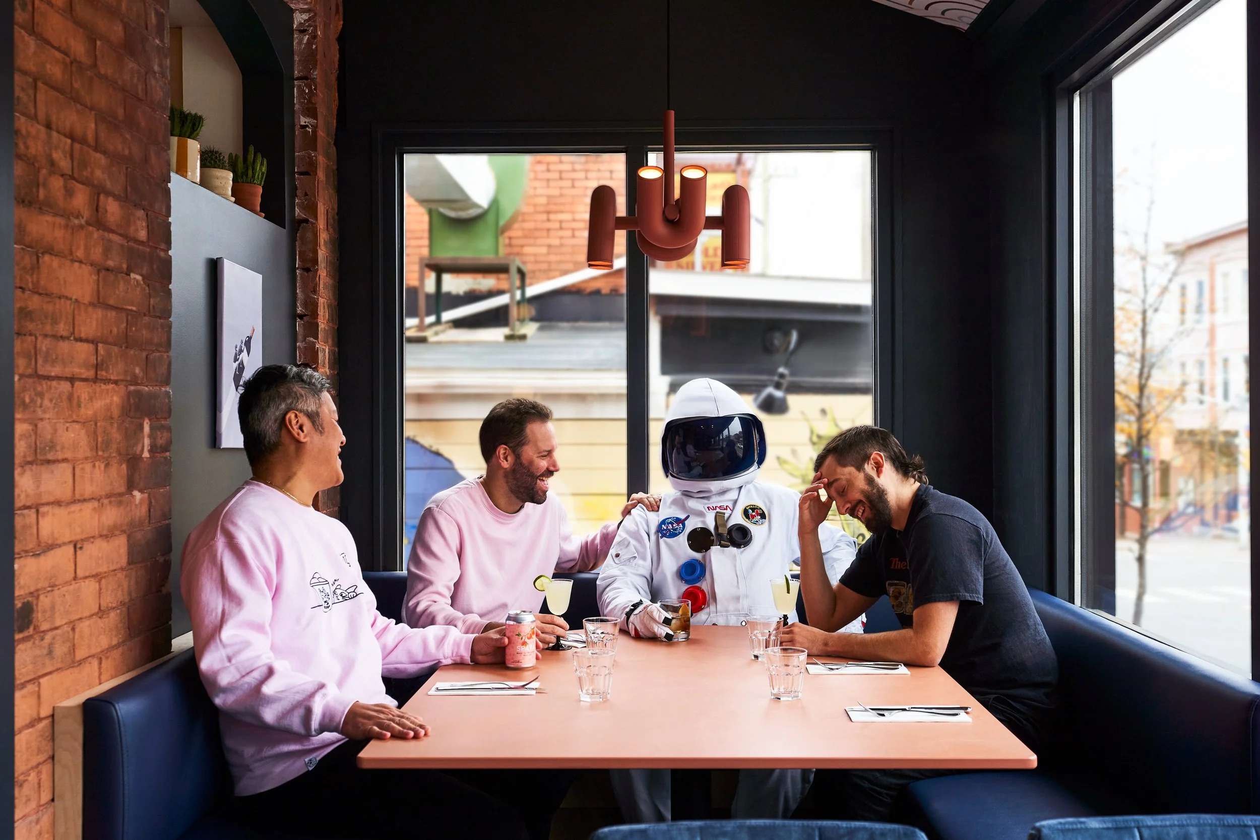 Four men are sitting at a wooden table in a restaurant with large windows, brick walls, and a modern ceiling light fixture. One of them is wearing a NASA astronaut suit. They are smiling and appear to be enjoying a friendly conversation.