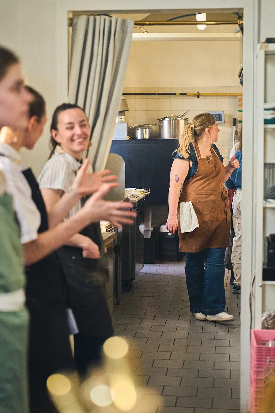 Kitchen staff waiting in line, with one woman in brown apron and others clapping and smiling, seen through a curtain in a restaurant kitchen. Photographed by Gumption Studio, Ottawa event photographer.