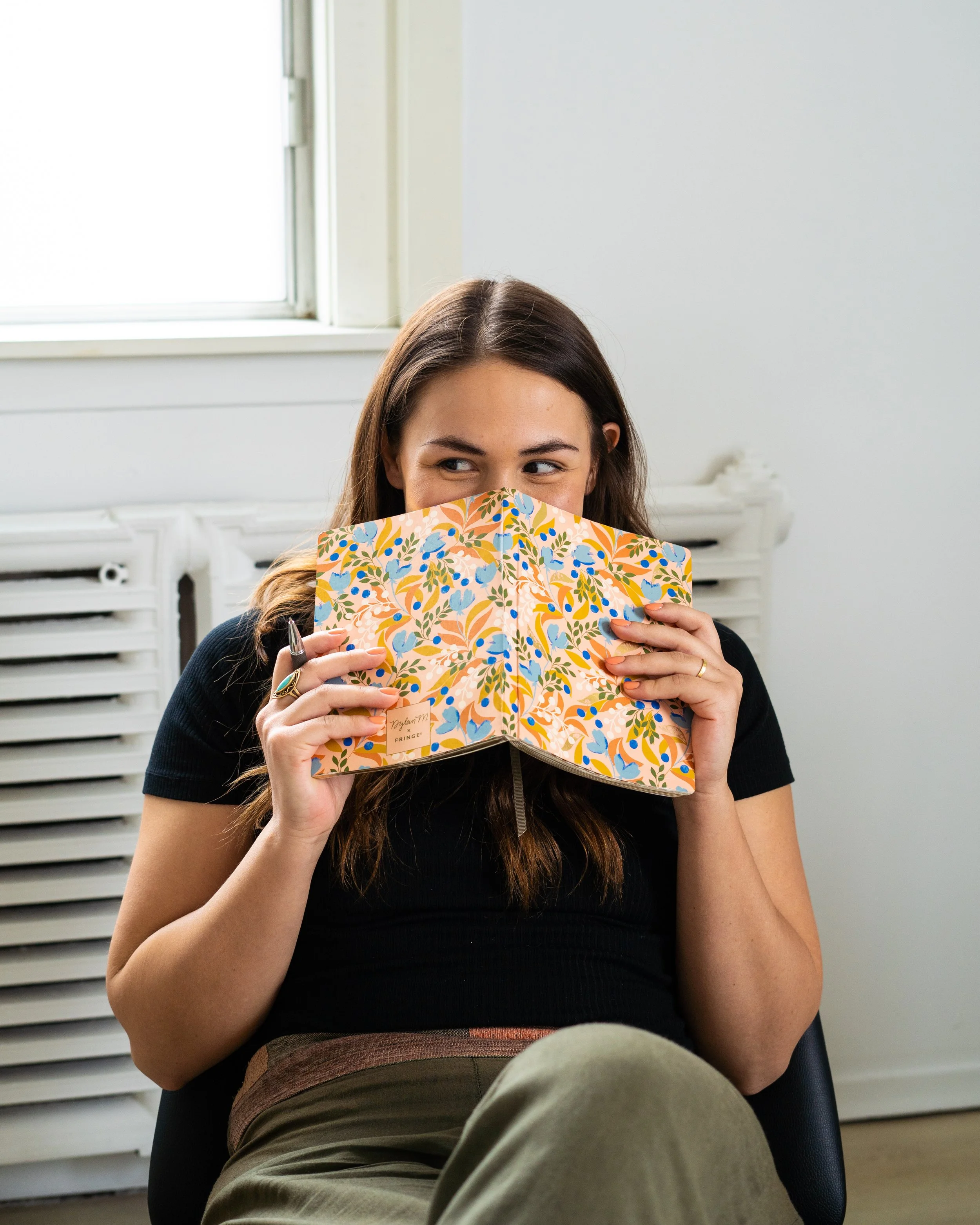 A woman with long dark hair is sitting in a chair indoors, partially hidden behind a colorful, patterned notebook or folder she is holding in front of her face. She is wearing a black top and appears to be looking to her right with a slight smile. Th