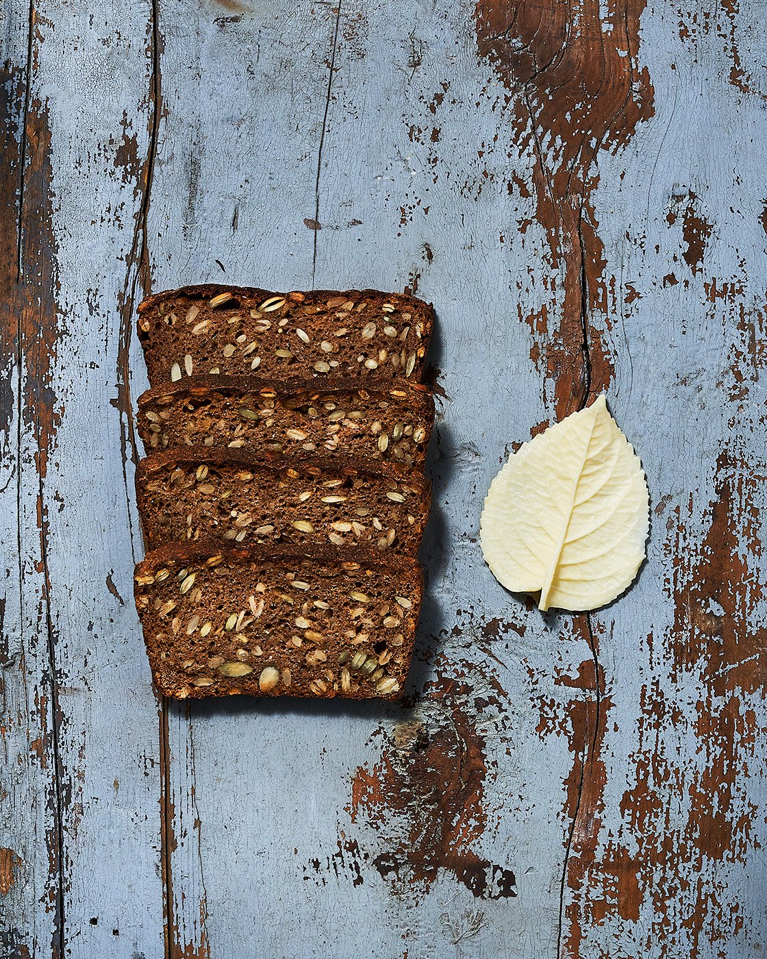 Four slices of sunflower seed bread next to a cluster of three white leaves of cultured butter on a distressed blue wooden surface. Photographed for the Perch Cookbook by Gumption Studio, Ottawa Cookbook photographer.