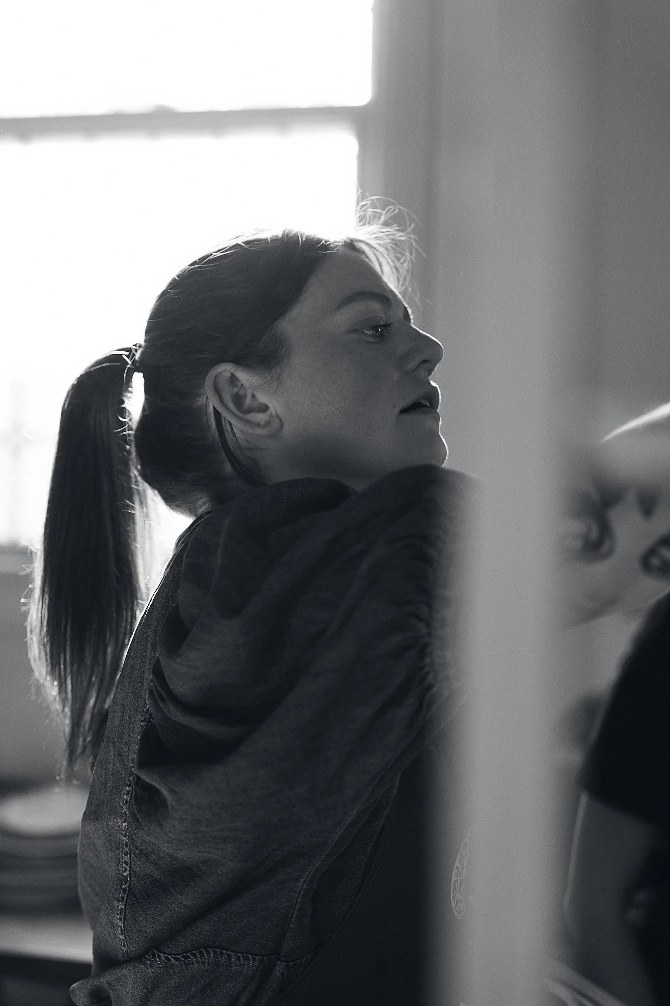 A young woman with long hair tied in a ponytail, wearing a hooded sweatshirt, leaning against a washing machine or dryer, looking to her right with a neutral expression. The background shows a brightly lit window and part of a laundry room.
