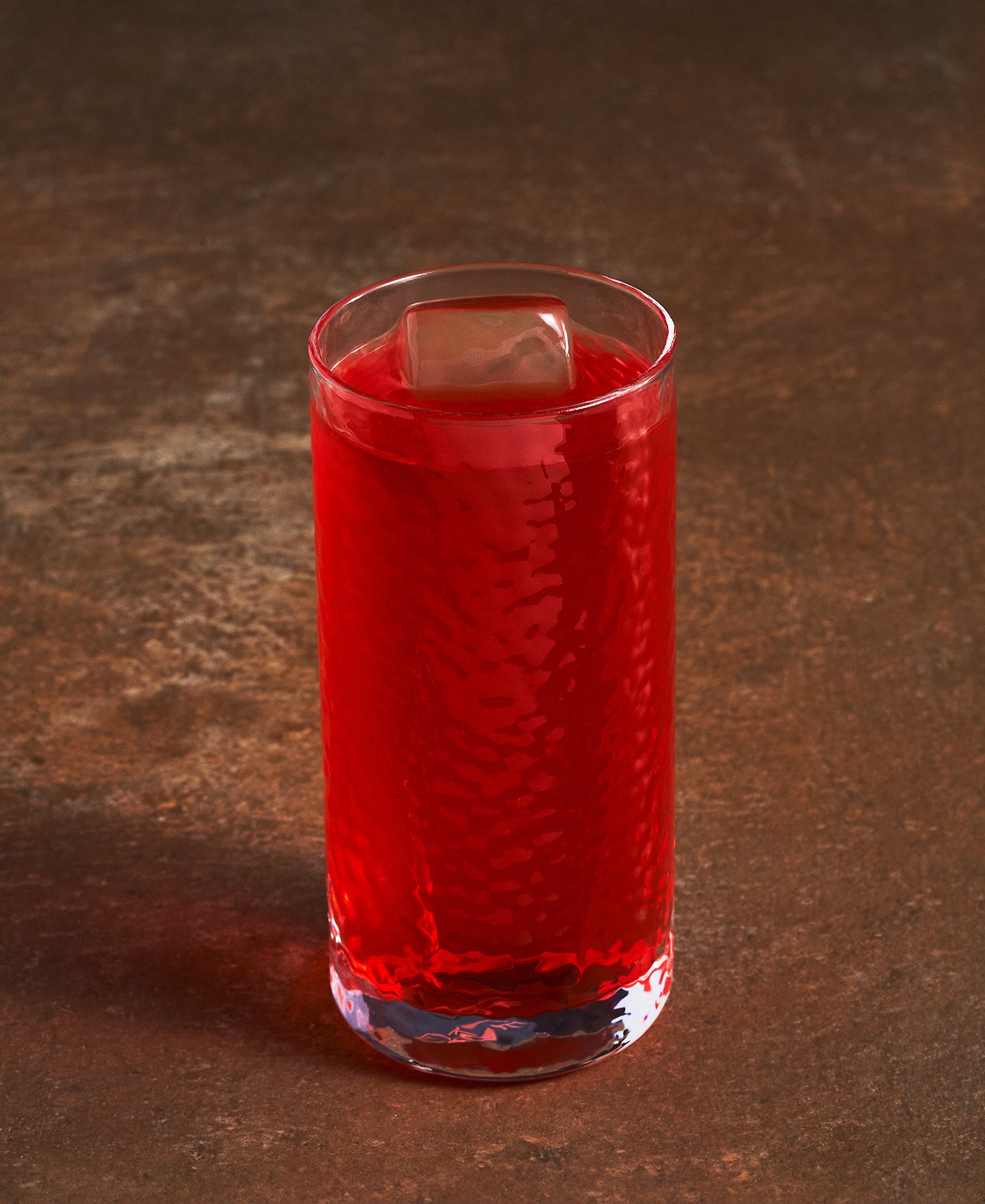 A tall, clear glass with a textured red surface, filled with a red beverage and a large ice cube, on a dark wooden surface. Photographed for the Perch Cookbook by Gumption Studio, Ottawa Cookbook photographer.
