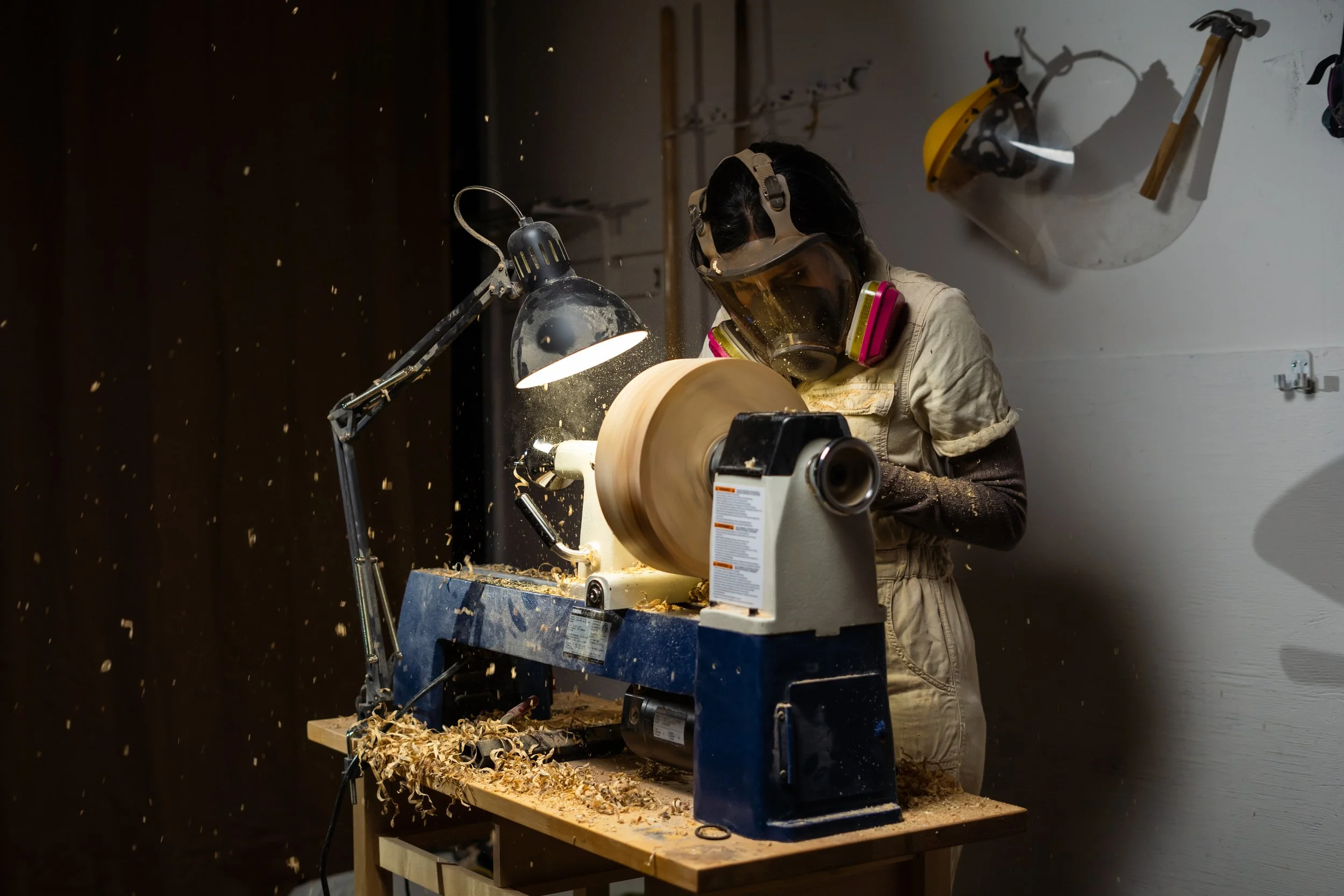 A person wearing safety goggles and a dust mask operates a woodworking lathe, shaping a piece of wood with wood shavings flying around. The work area has a lamp illuminating the workspace, and tools are mounted on a white wall behind.