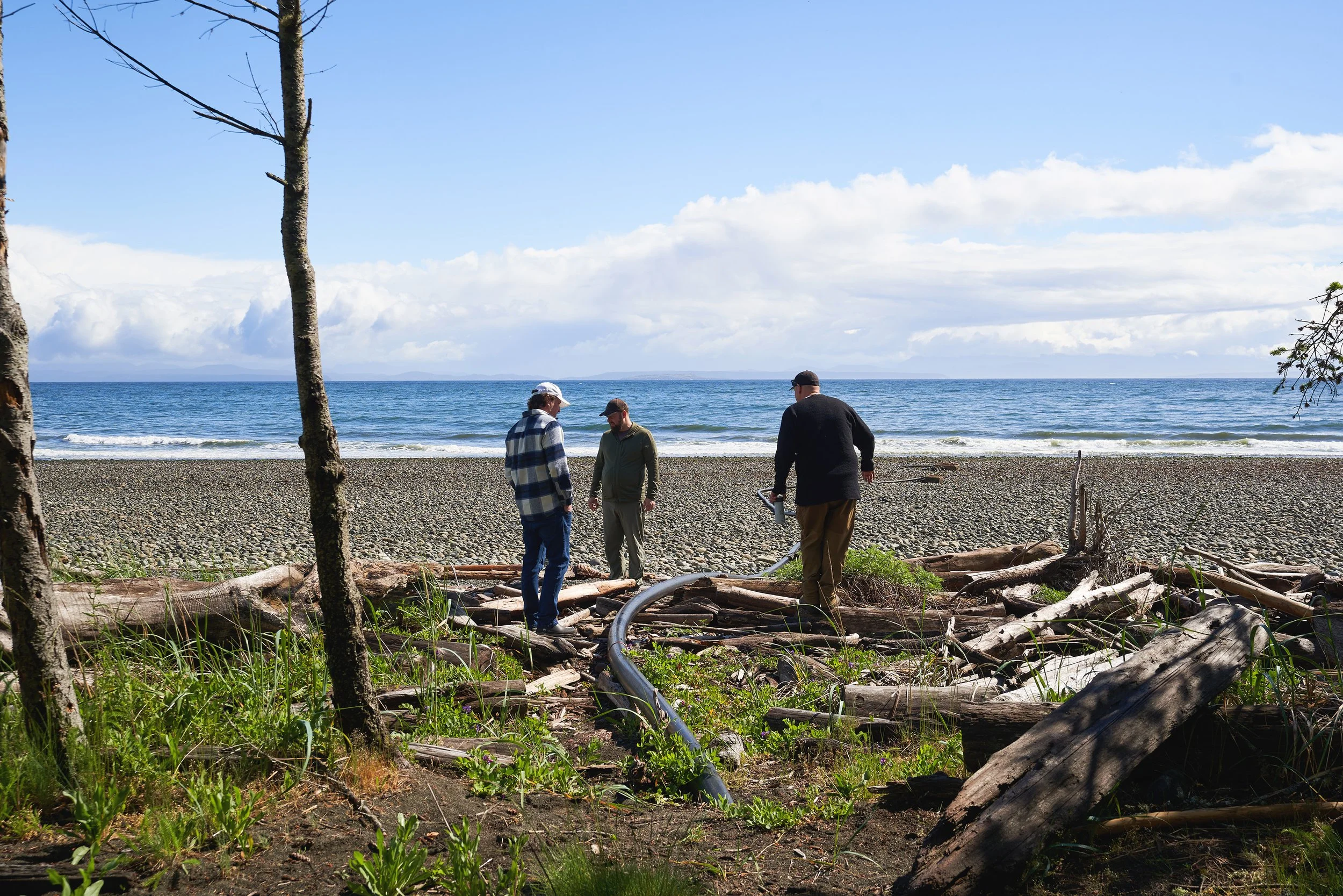 Three men standing on a pebble beach at Vancouver Island Sea Salt, with trees and driftwood in the foreground, and a blue sky with clouds overhead. Photographed for the Perch Cookbook by Gumption Studio, Ottawa Cookbook photographer.
