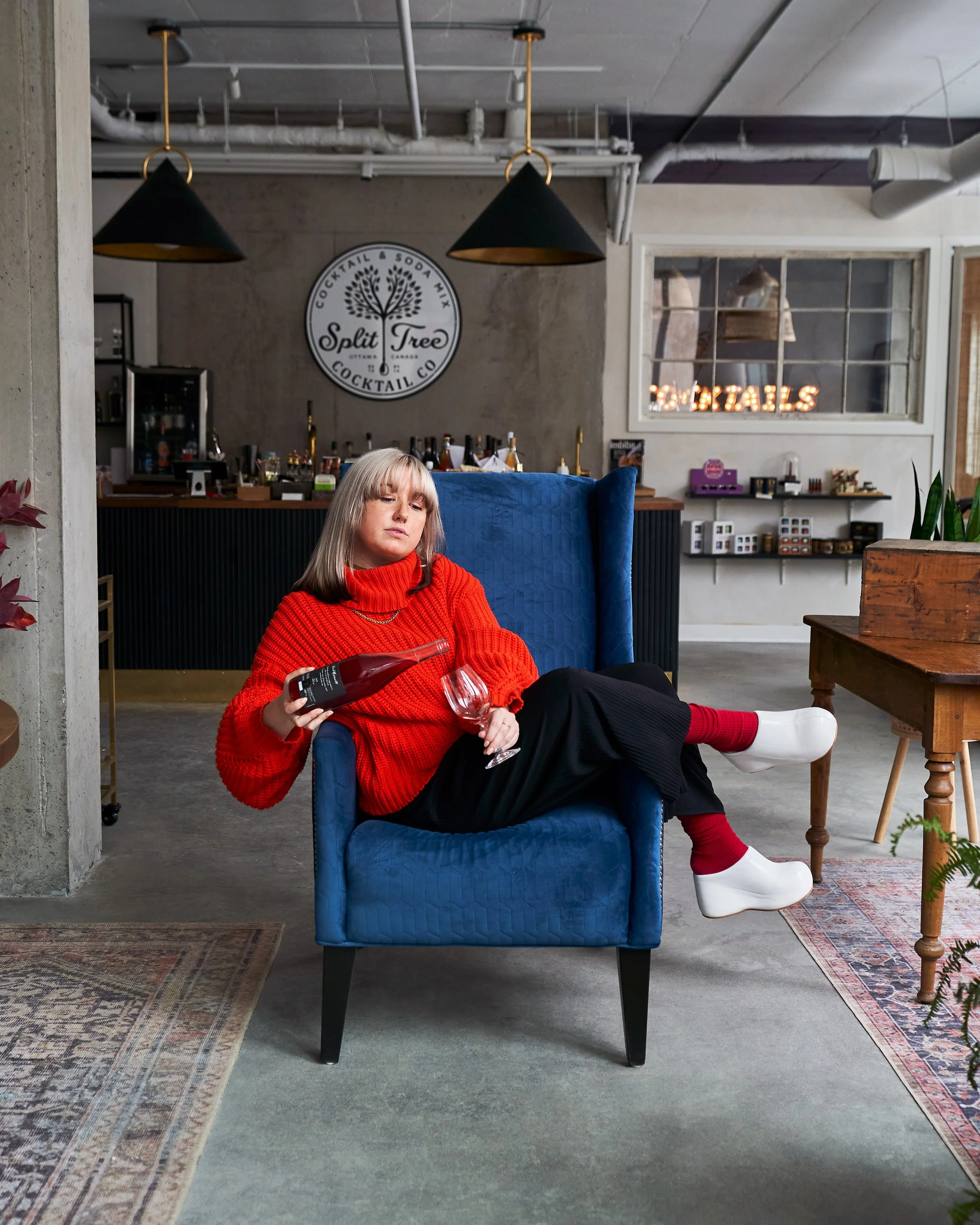 A woman with long blonde hair wearing a bright red sweater and black pants, seated in a blue armchair, holding a bottle of wine in one hand and a wine glass in the other, inside a modern lounge area with industrial style decor, including pendant ligh
